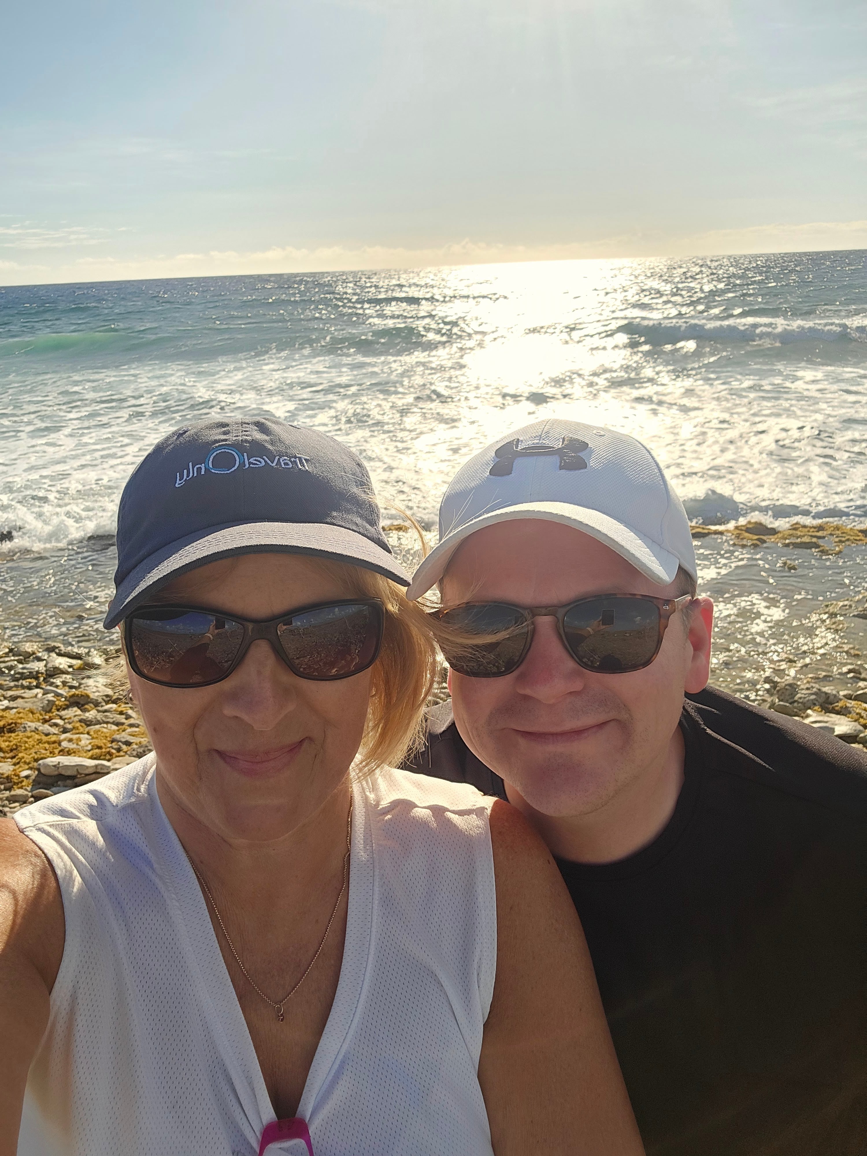 Two people in casual beachwear posing on rocky Aruba shoreline with turquoise ocean behind