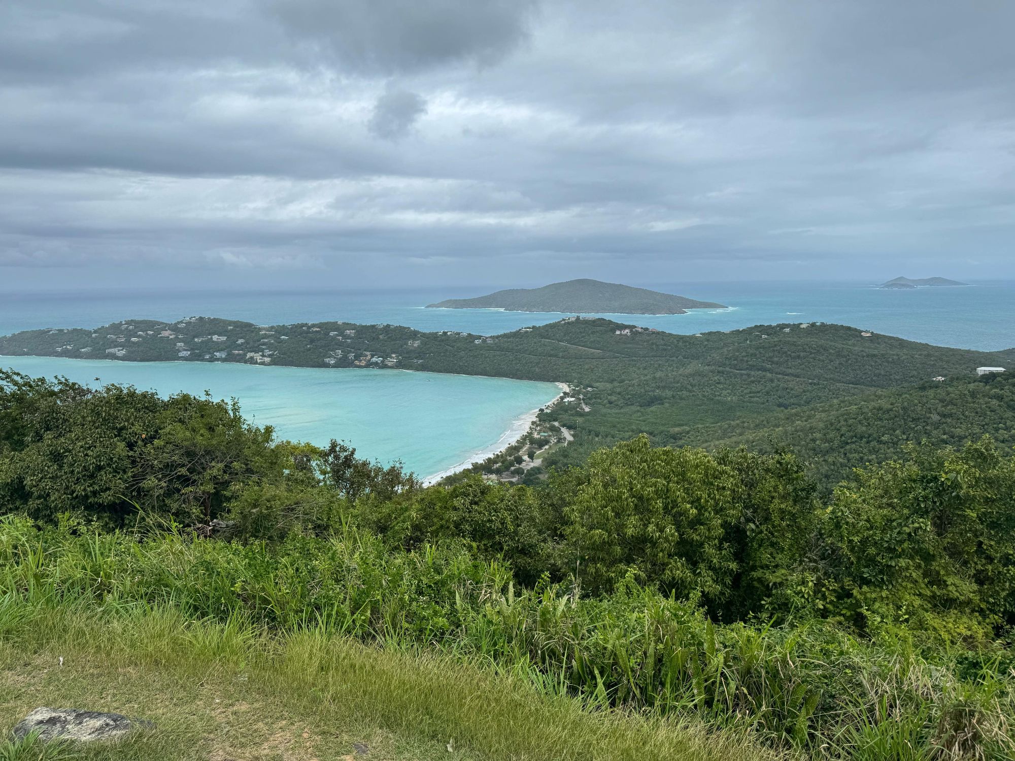A sweeping aerial view of a turquoise Caribbean bay nestled between lush green hills under stormy skies.