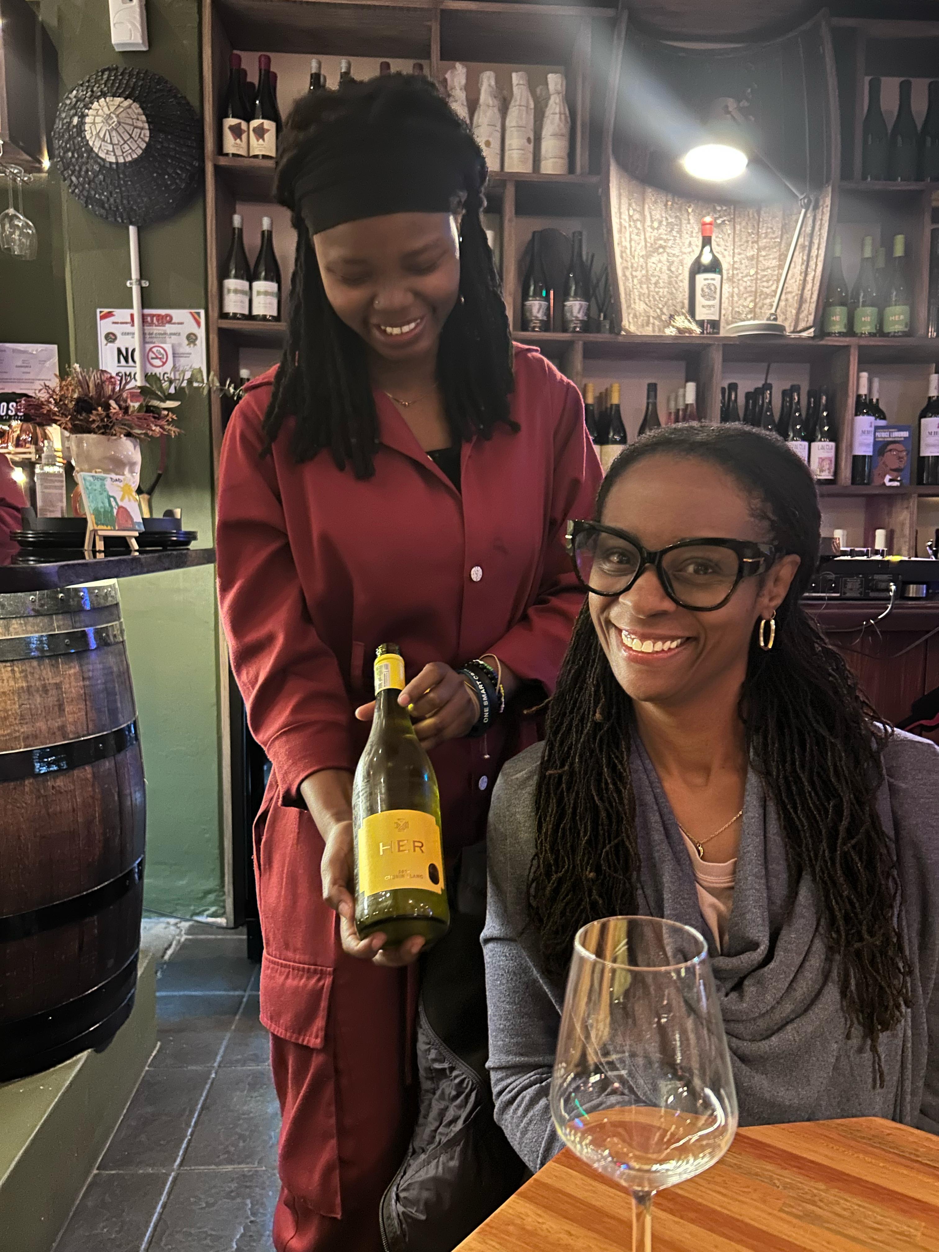 Two women at a wine tasting, one in burgundy holding a yellow wine bottle, shelves of wine bottles visible behind
