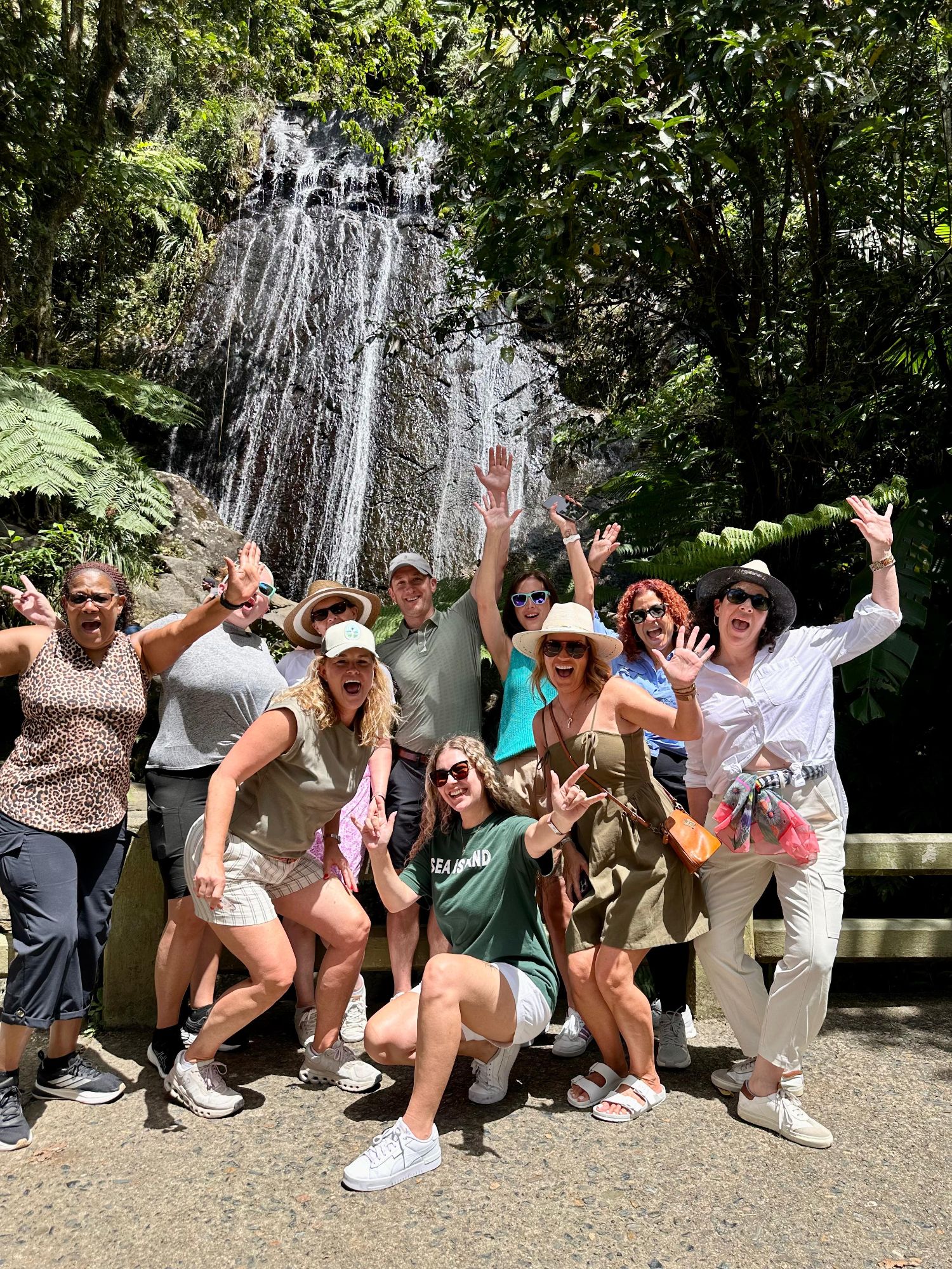 Large group posing enthusiastically in front of a waterfall surrounded by lush green forest