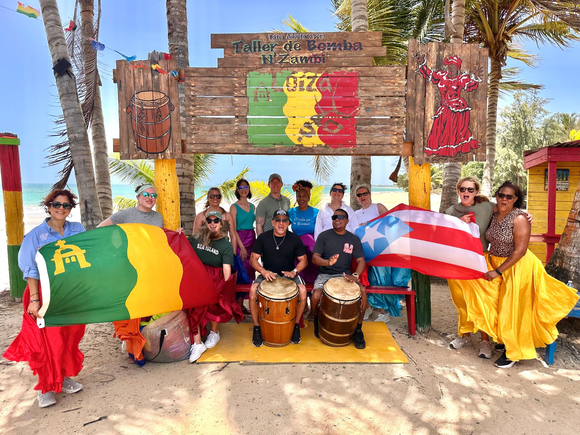 Group holding Cameroon and Puerto Rico flags at a beach drum circle with colorful wooden structures