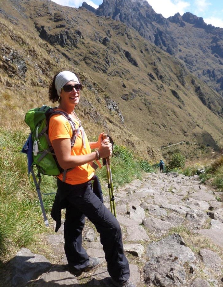 Tannis Dyrland hikes on a rocky mountain trail wearing an orange shirt and green backpack with dramatic peaks in the background