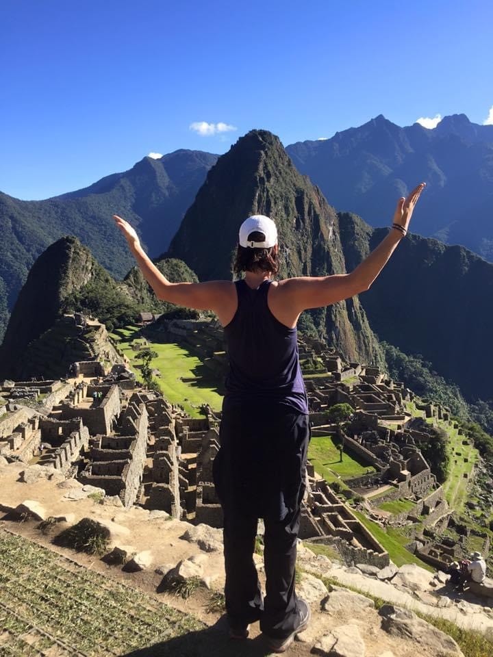 Tannis Dyrland stands with arms raised at Machu Picchu, with ancient stone structures and green mountains visible behind her