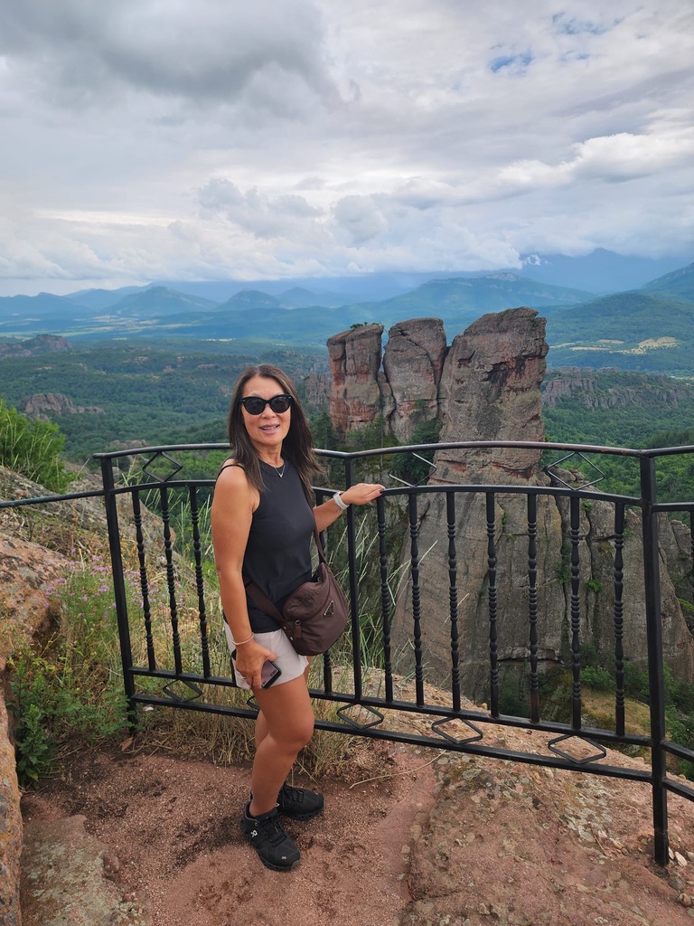 Grace Wong poses at a viewpoint railing with distinctive rock formations and a vast green valley landscape behind her