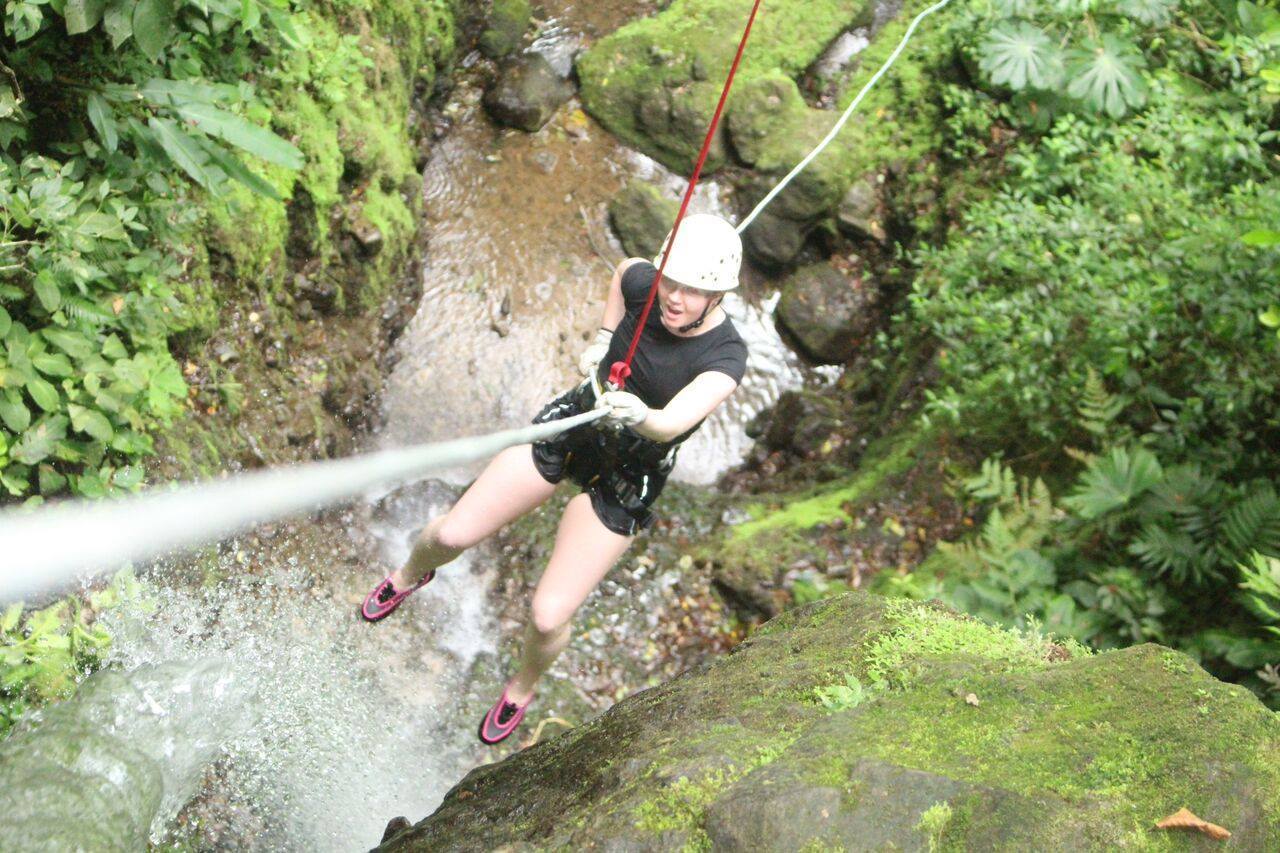 Autumn Fiske rappels down a moss-covered canyon wall on a red rope, suspended above a stream in Costa Rica