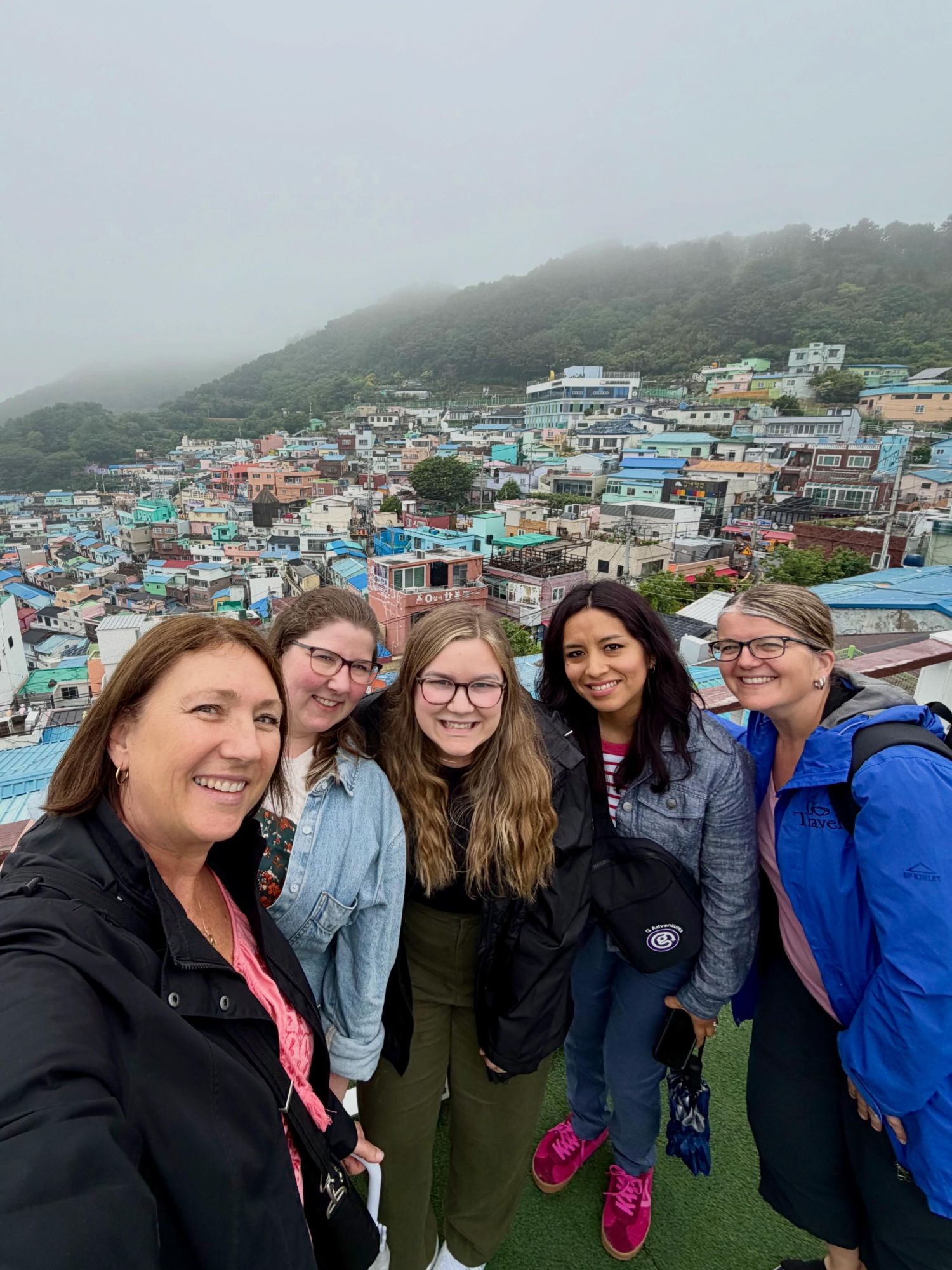 Five women smiling at a viewpoint overlooking a colorful hillside village with misty mountains