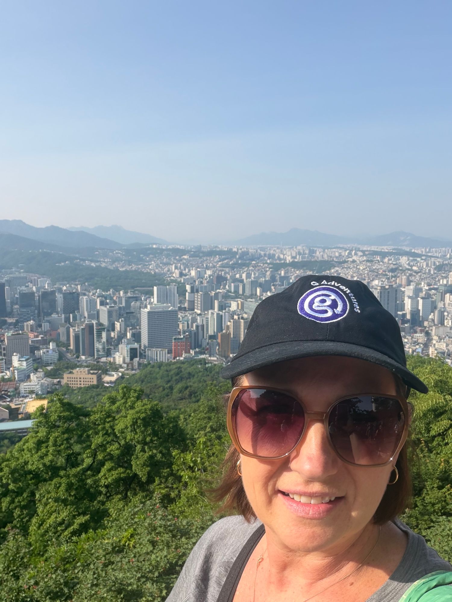 Woman wearing a cap and sunglasses with a panoramic view of Seoul's cityscape and surrounding mountains