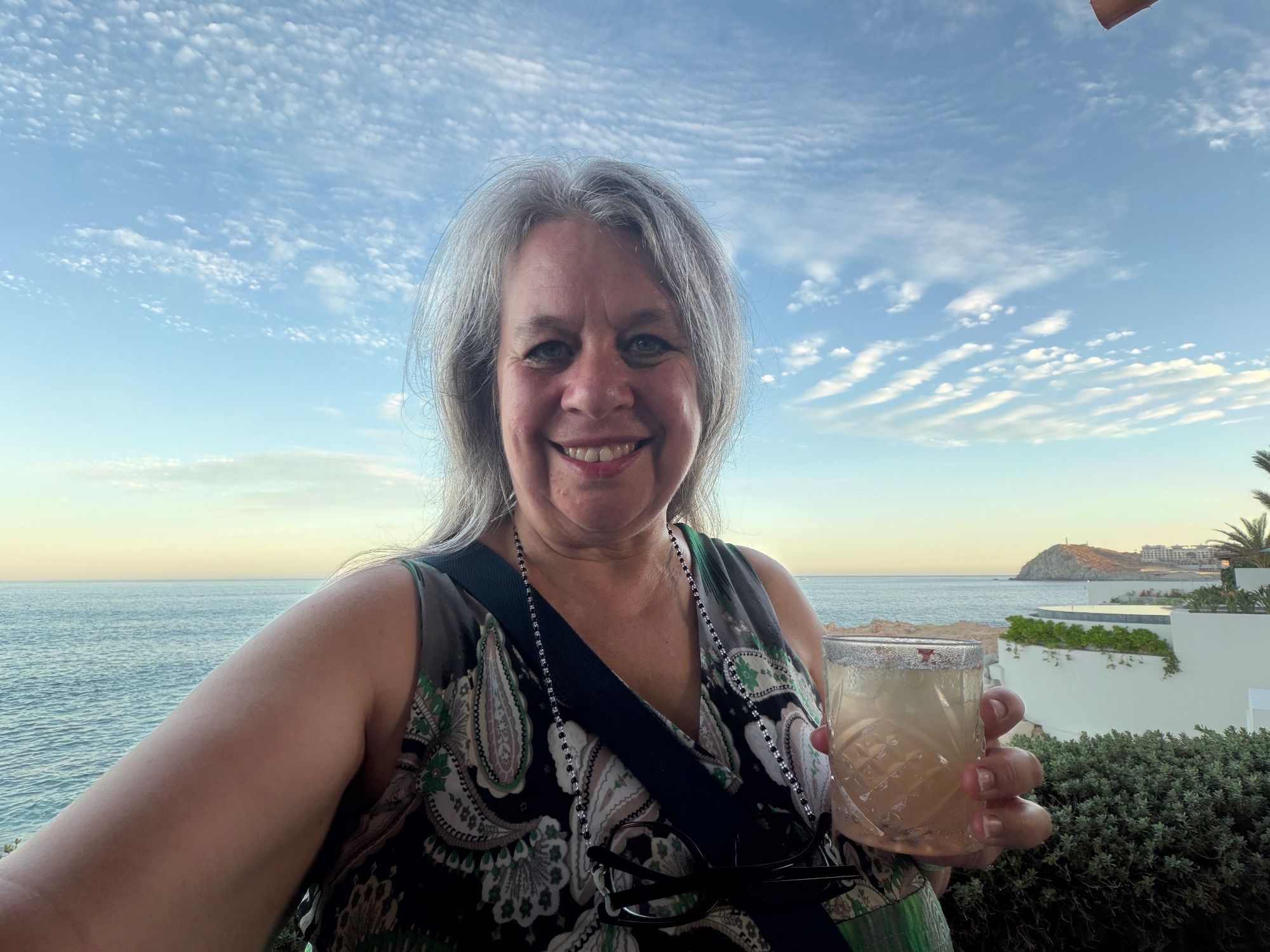 Woman smiling at sunset on a coastal terrace holding a drink with calm ocean and rocky islands visible behind her