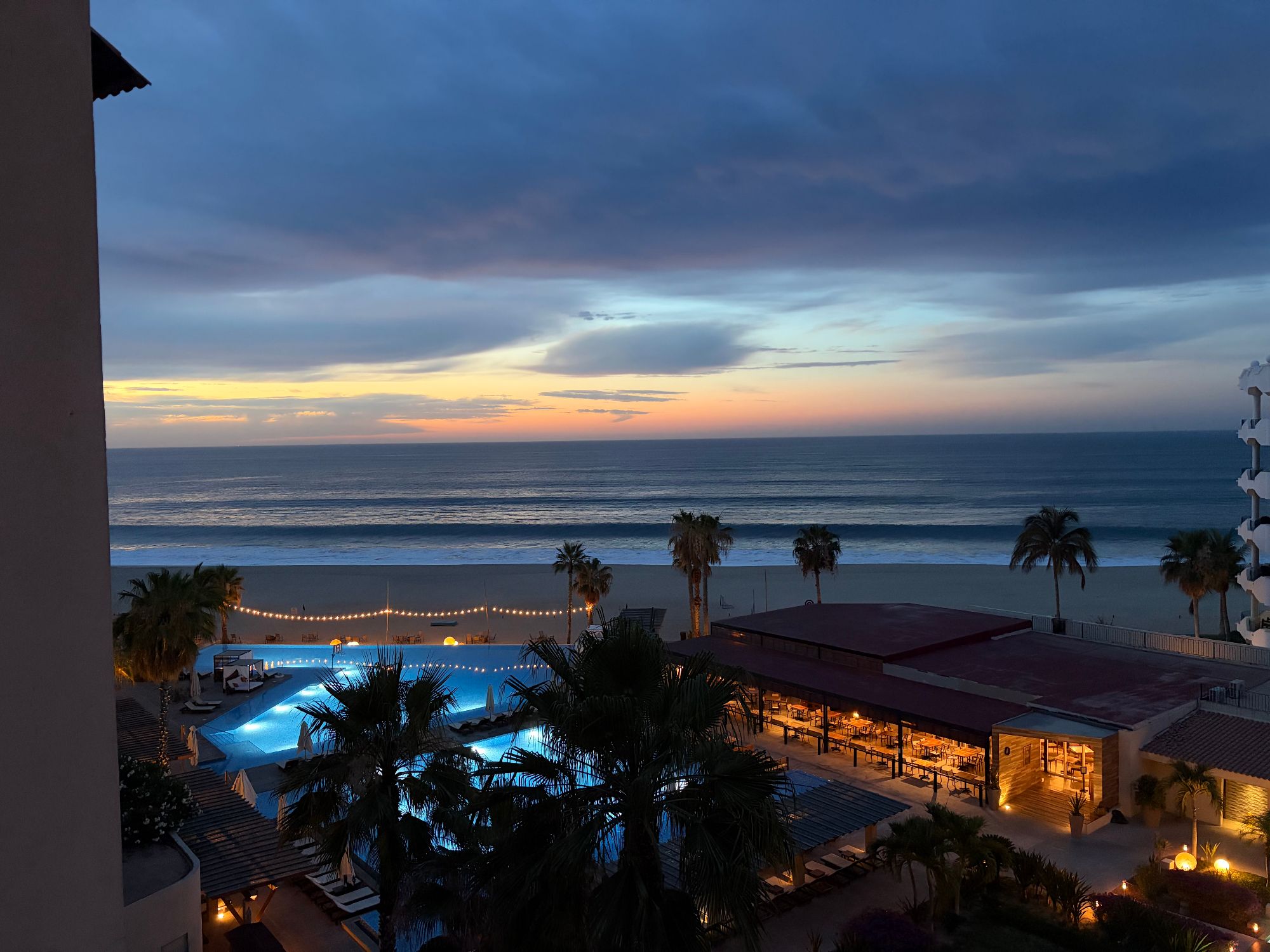 Beachfront resort at dusk with illuminated pool, palm trees, and ocean horizon during golden hour sunset