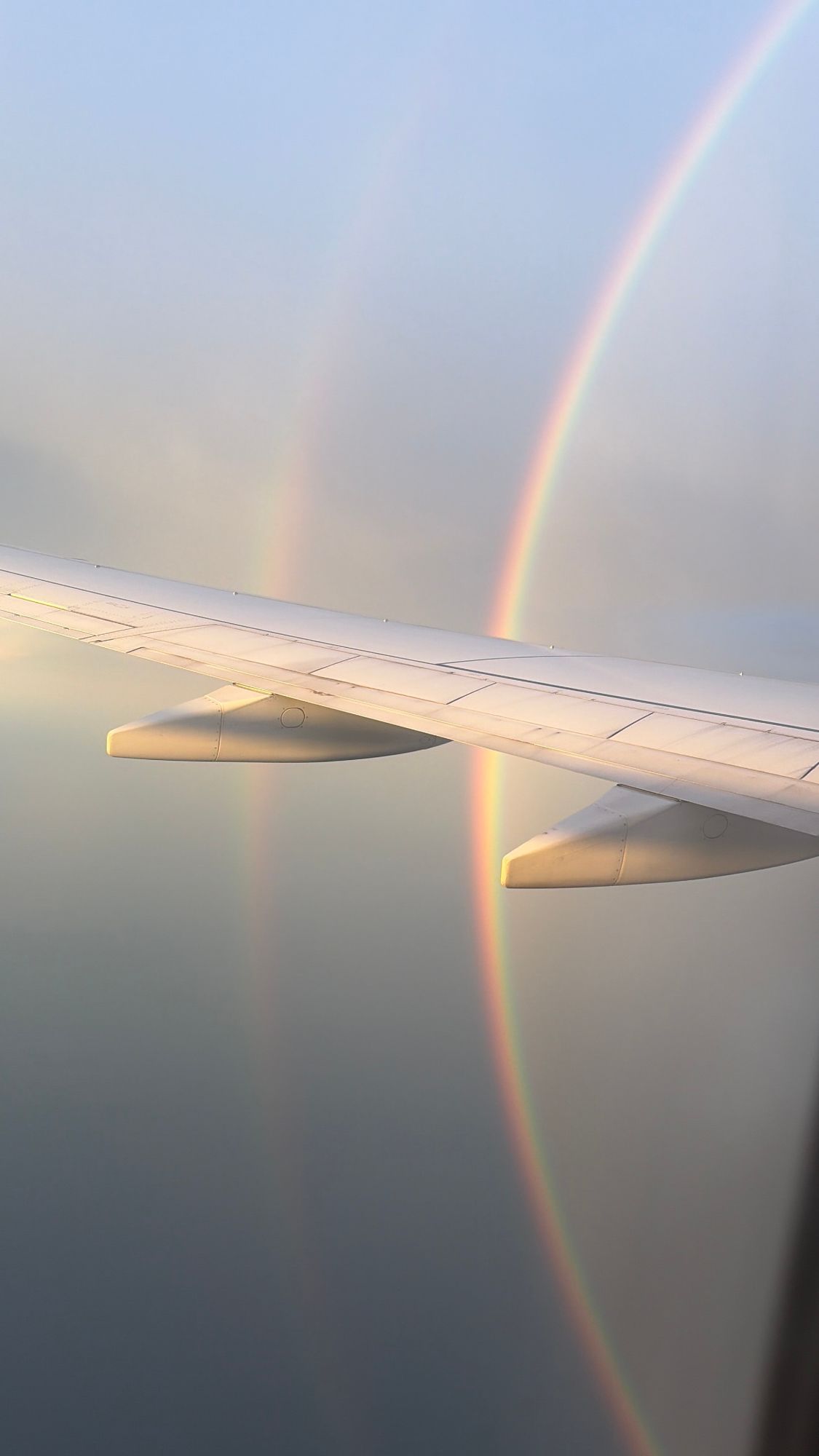 Rainbow visible from airplane window with aircraft wing and engines illuminated by golden sunlight