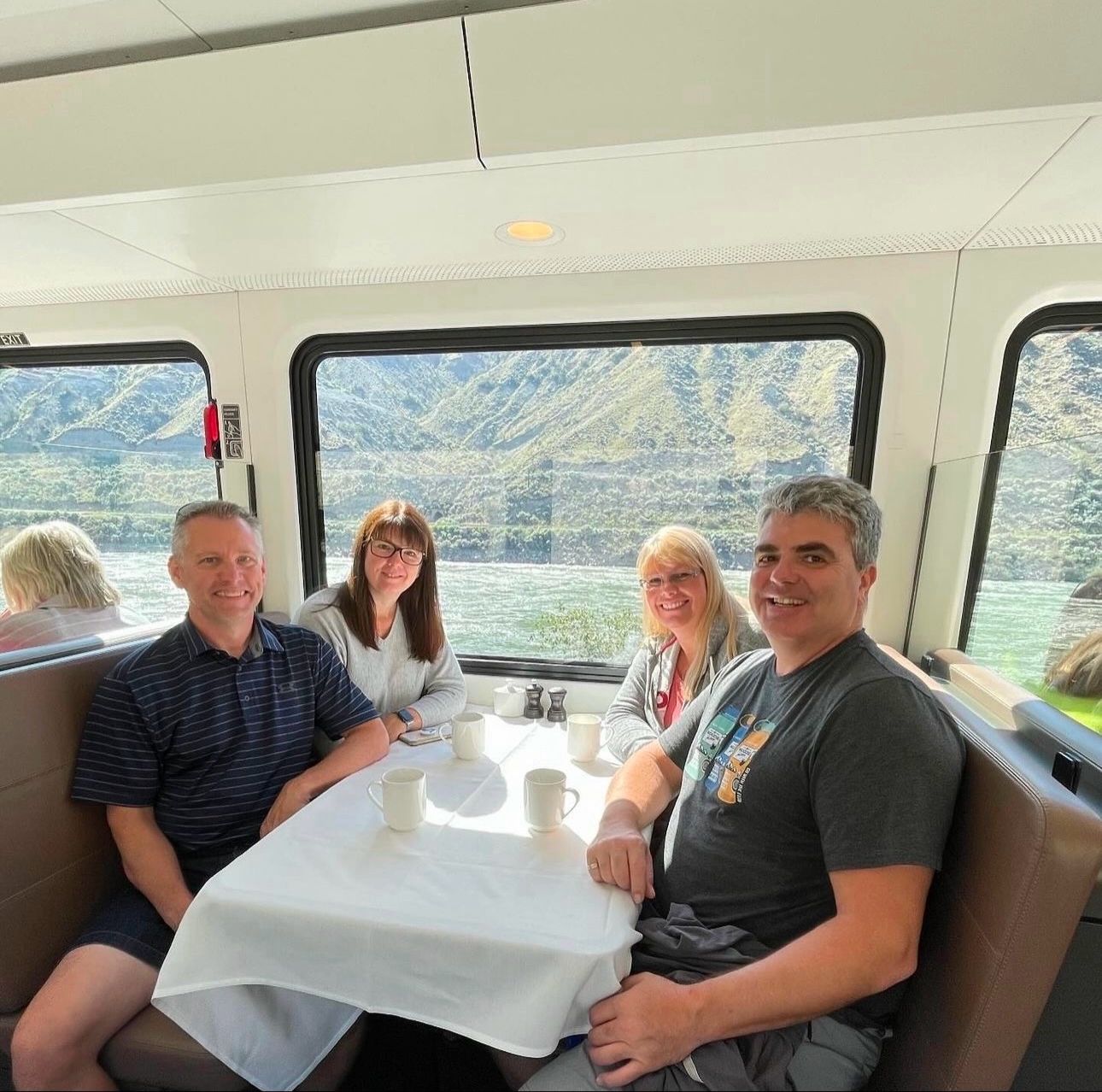 Four passengers dining at a table inside a modern train car with mountain and river views through large windows