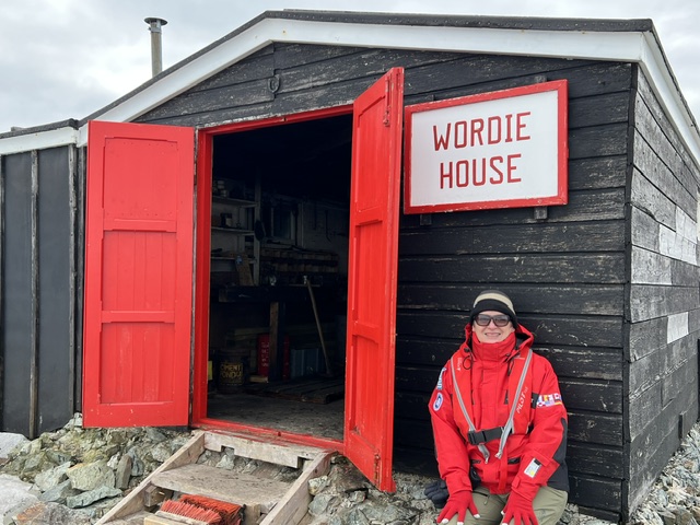 Theresa Bobrowsky stands in red cold-weather gear outside the Wordie House, a black wooden structure with red doors