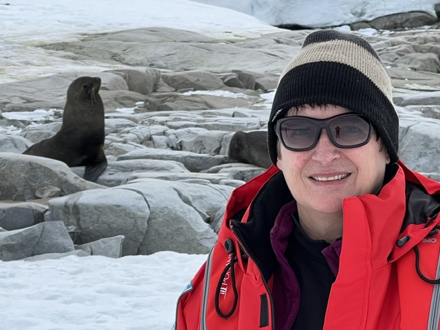 Theresa Bobrowsky smiles at the camera wearing red expedition gear and a striped beanie, with a seal visible on ice behind her