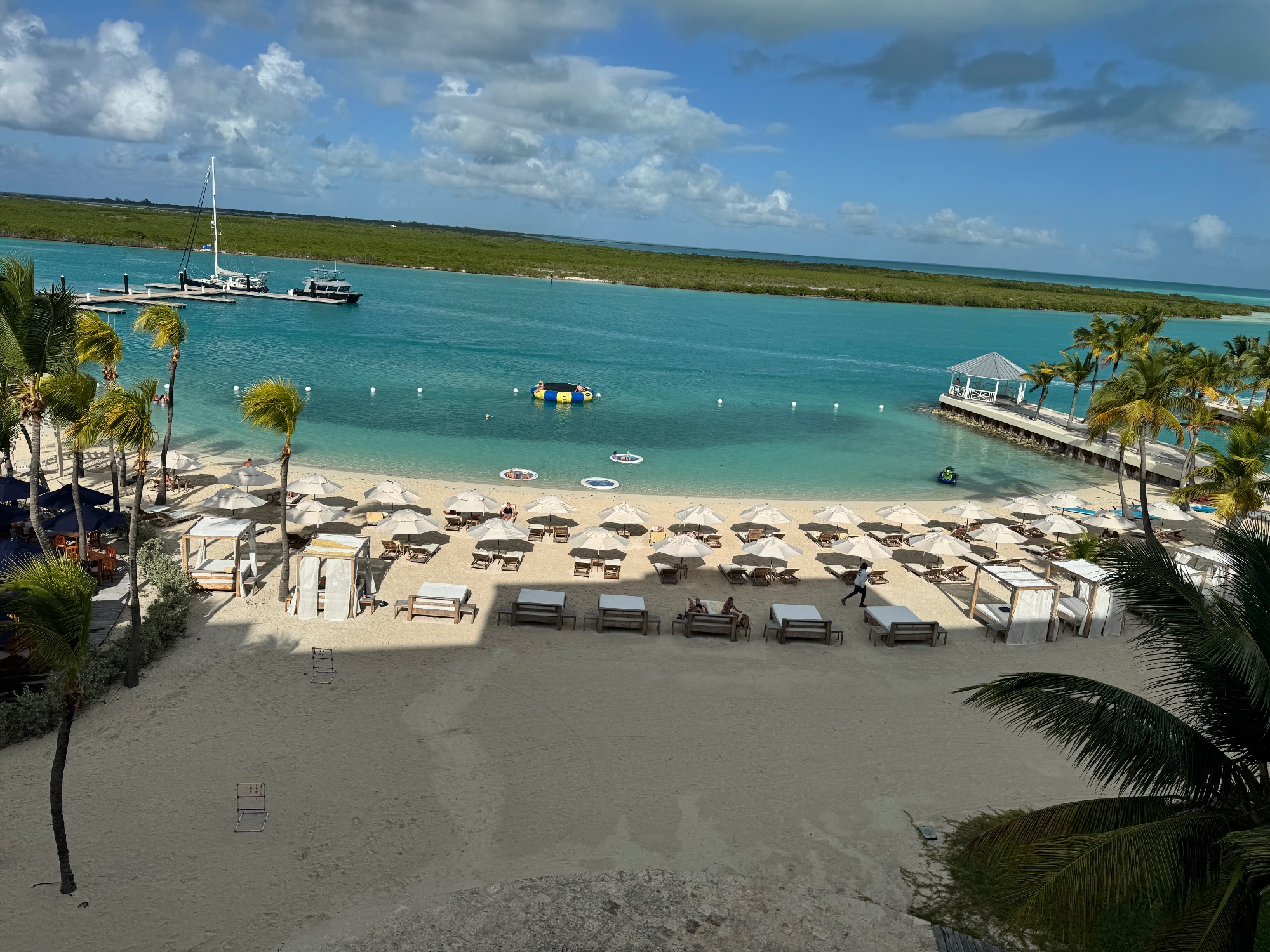 Elevated view of a sandy beach with white umbrellas, lounge chairs, palm trees, and boats in crystal-clear turquoise water