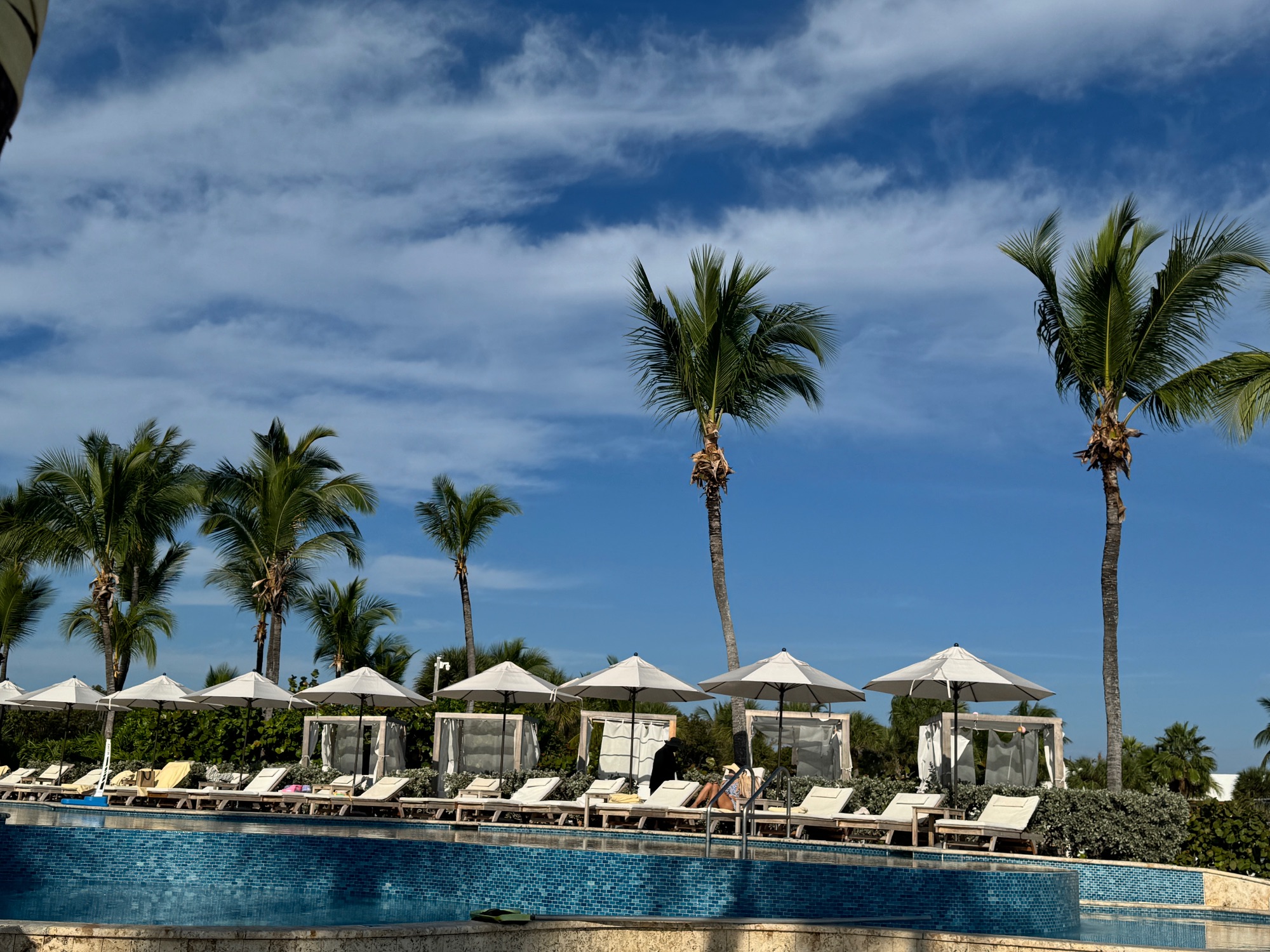 Beach cabanas with white curtains and palm trees overlooking a calm turquoise bay with moored boats
