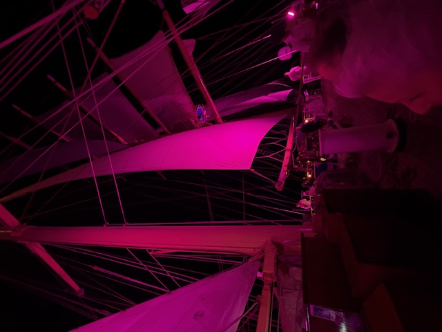 Interior of a ship's deck illuminated in magenta and pink lighting, showing the rigging and masts from below