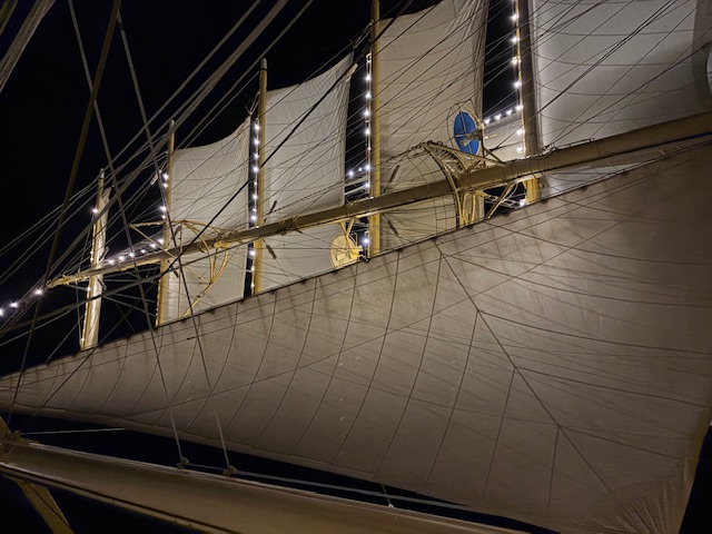 Illuminated white sails and rigging of a tall ship at night, with deck lights creating a dramatic upward view