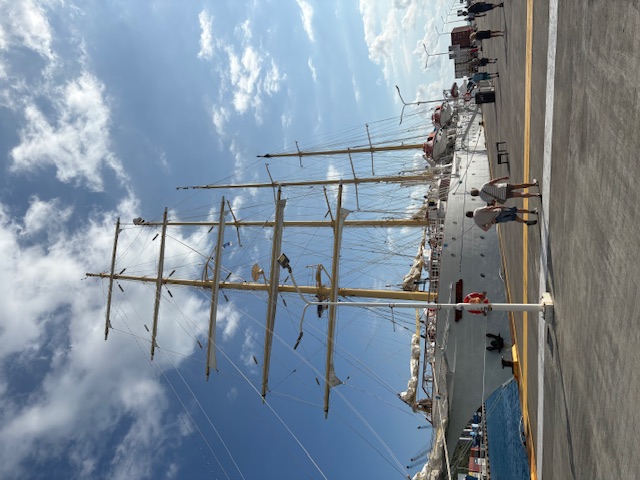 Four-masted tall ship docked at a waterfront with people walking on the pier and blue water alongside