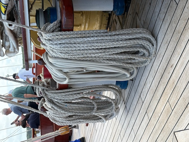 Coiled gray and white rope on the wooden deck of a sailing vessel with crew members visible in background