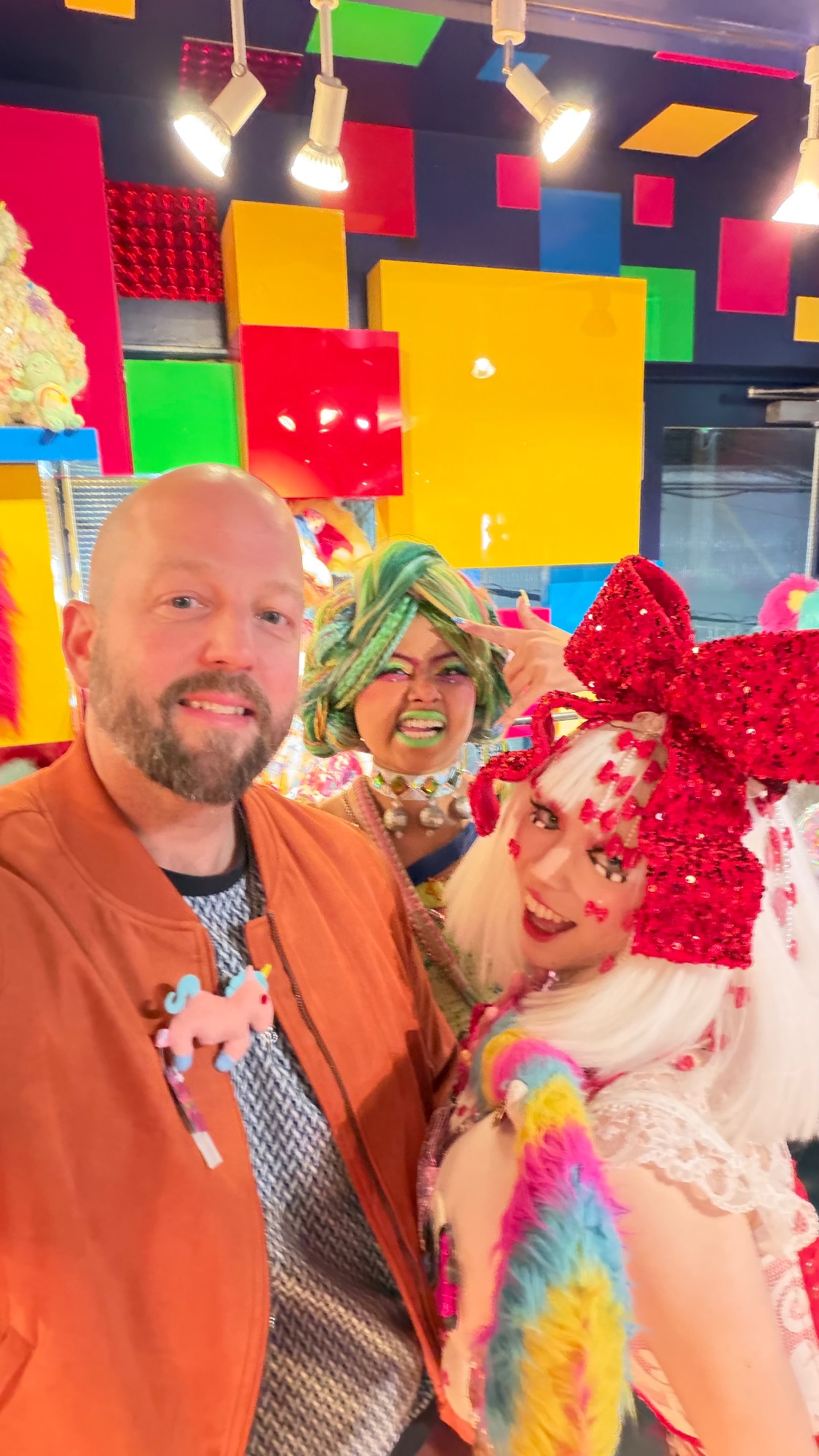 Duncan Greenfield-Turk smiles with two performers in colorful wigs and sequined outfits against a vibrant backdrop of primary-colored geometric panels
