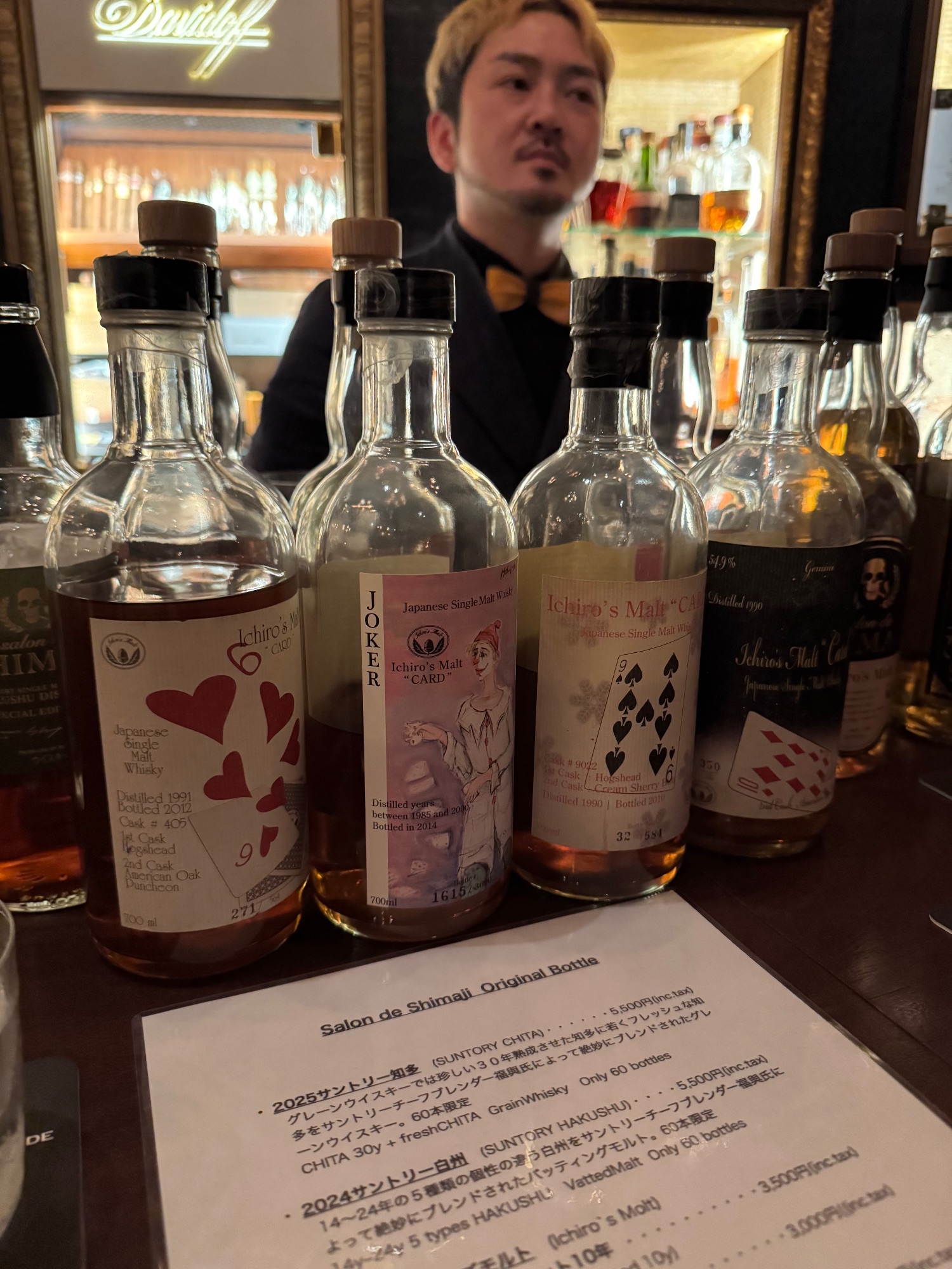 Japanese whisky bottles with playing card-themed labels displayed on a bar counter, with a bartender visible in the background