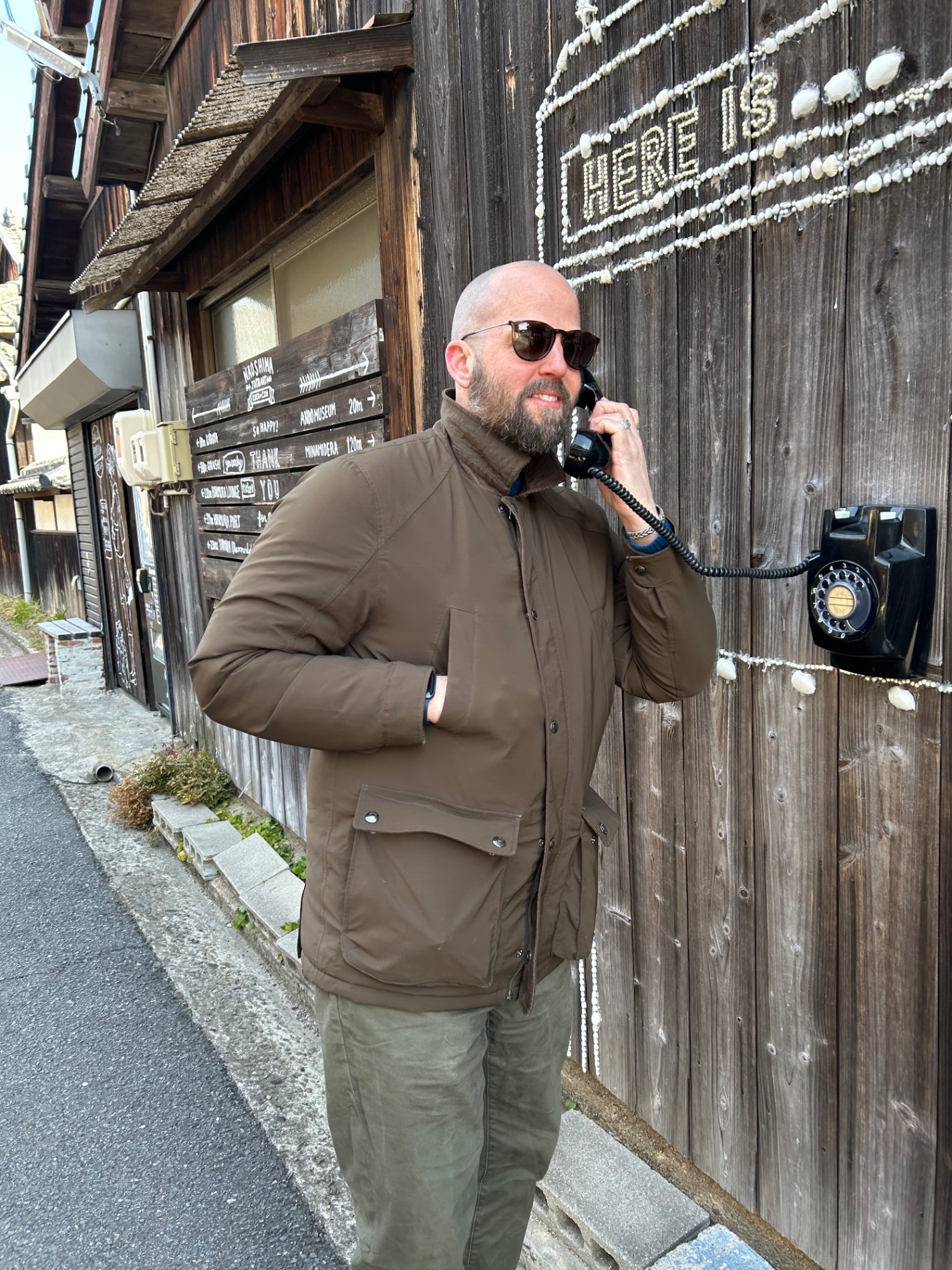 Duncan Greenfield-Turk speaks on a vintage rotary telephone outside a weathered wooden Japanese building with hand-painted signage