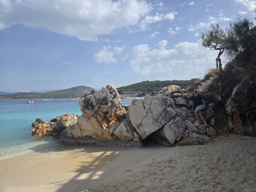 Rocky beach cove with large boulders and turquoise water, person standing on rocks with forested hillside in background