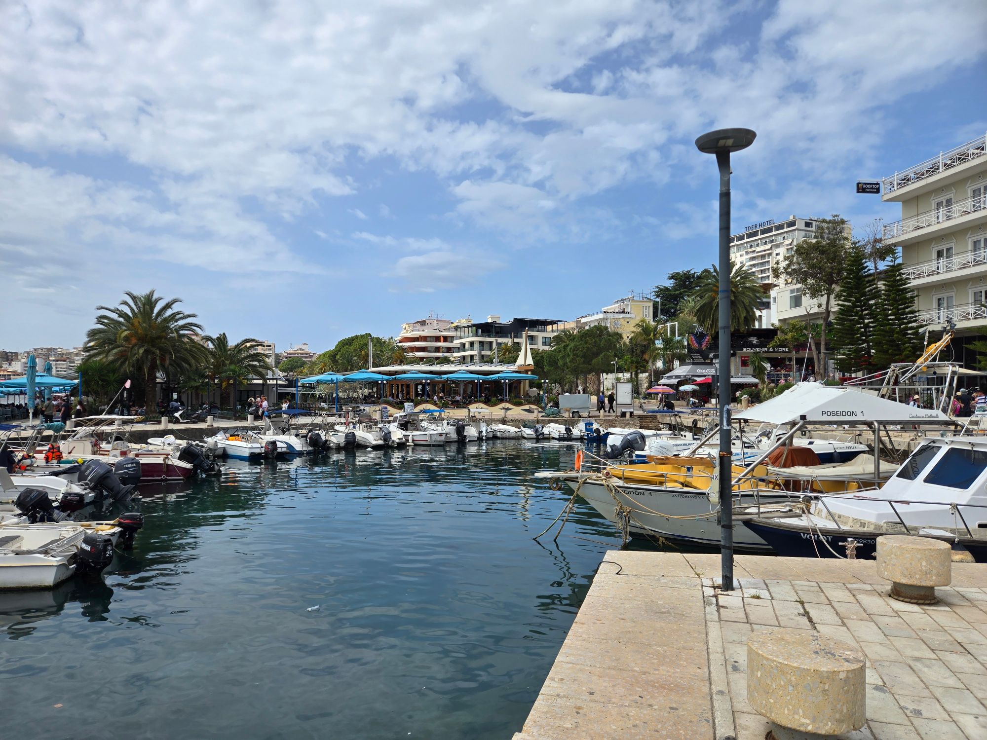 Busy harbor filled with moored fishing boats and yachts, waterfront cafes and palm trees lining the dock