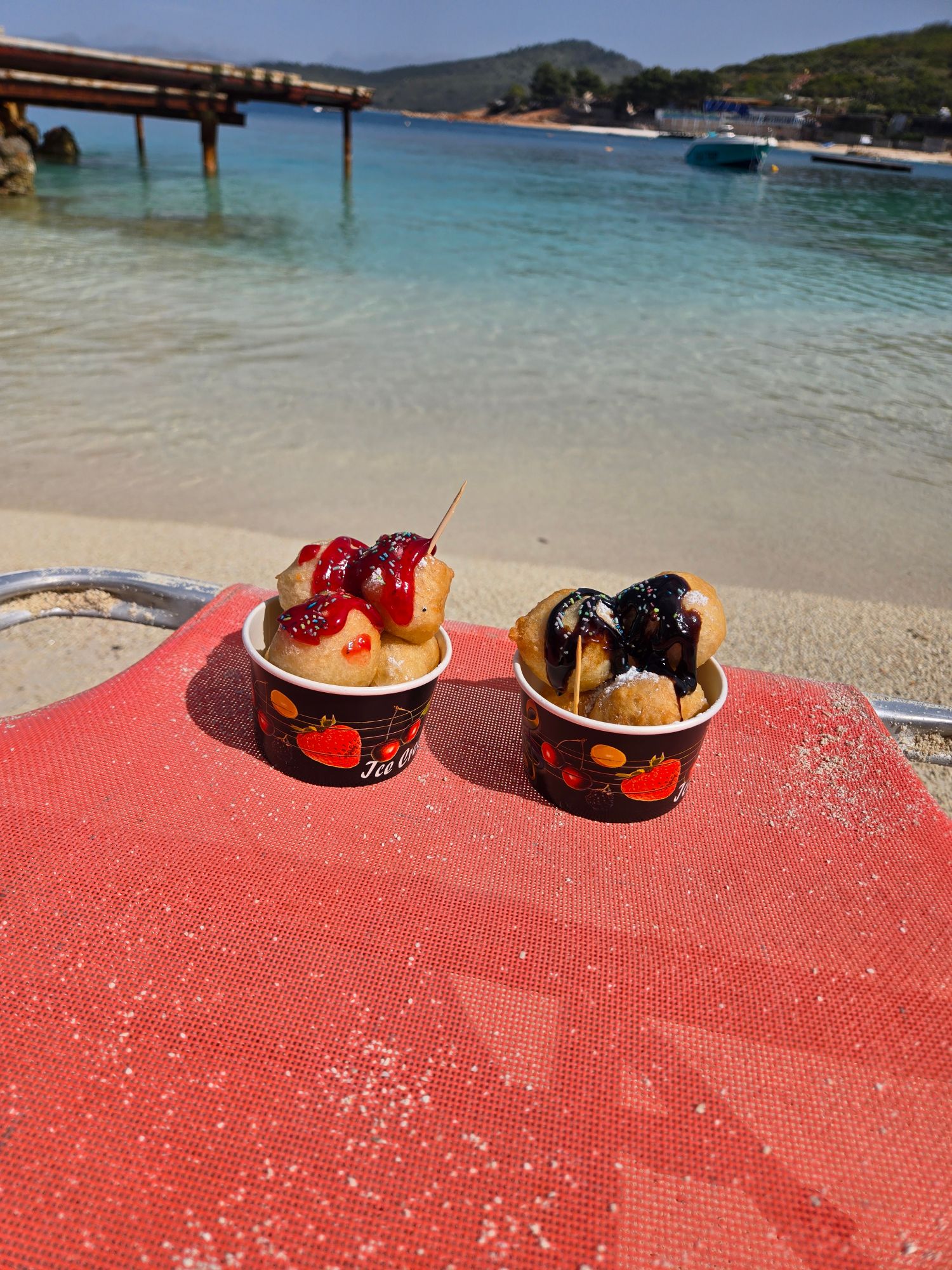 Two cups of fried dough desserts topped with red and dark sauce on a red mat overlooking a beach and moored boats