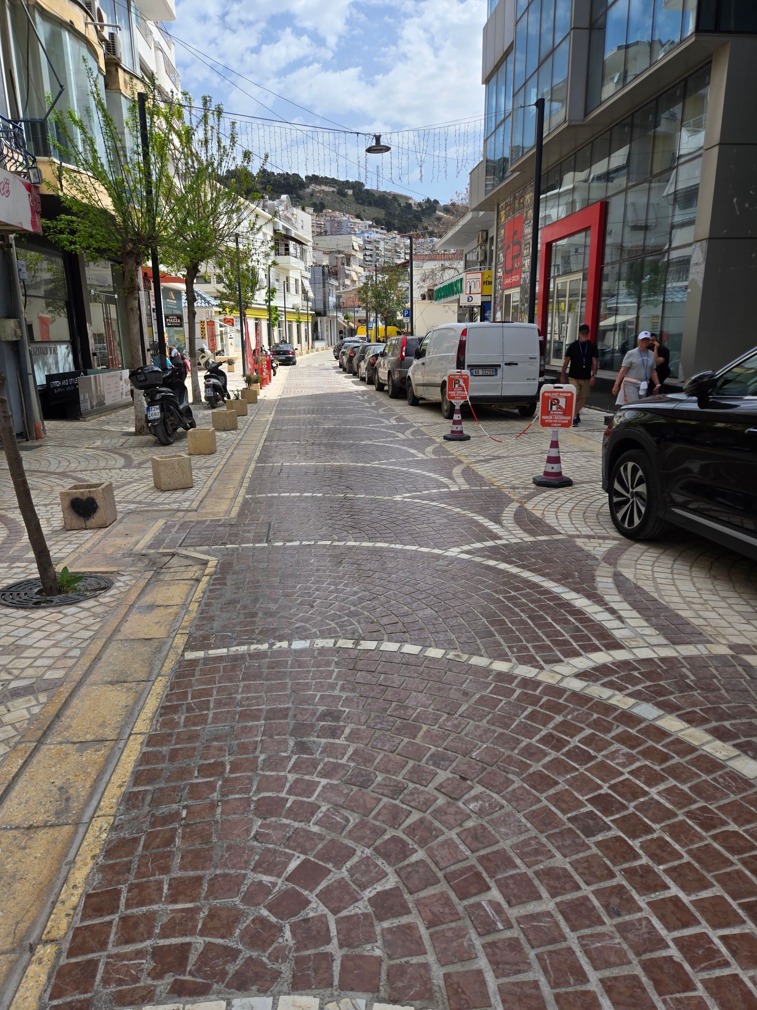 Pedestrian shopping street lined with shops and parked cars, hillside with buildings visible at the end of the street