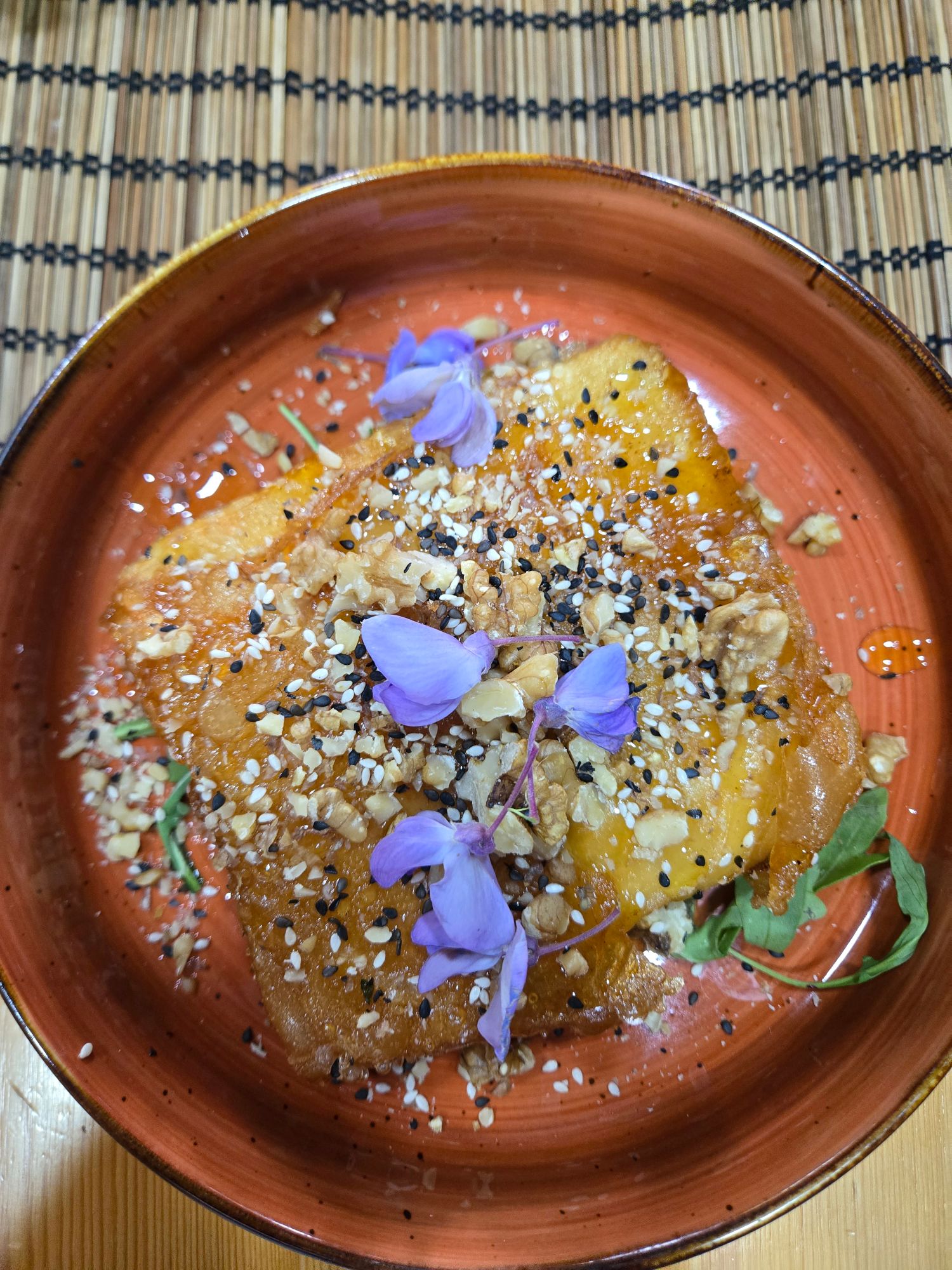 Plate of hummus or similar spread topped with nuts, seeds, and purple flowers on a woven placemat