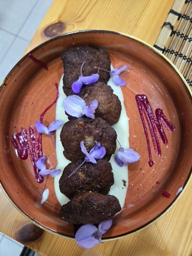 Plate of three chocolate truffles or meatballs with white cream, purple flowers, and dark sauce drizzle on terracotta dish