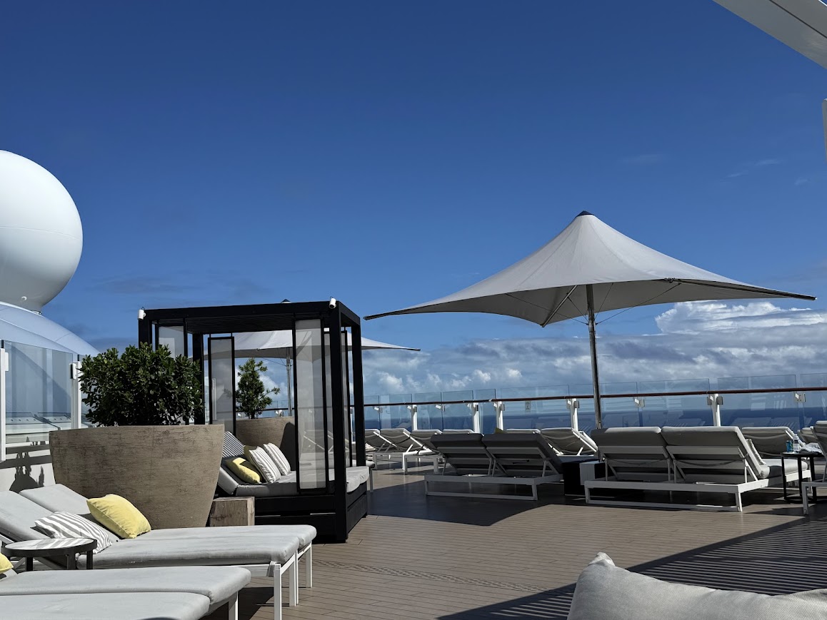 Ship's upper deck lounge area with modern furniture, umbrellas, and expansive ocean view under blue sky