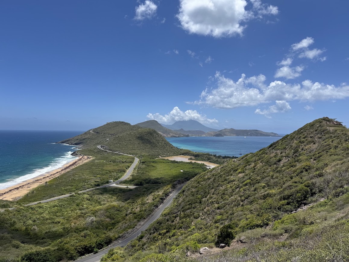 Lush green hillside landscape with winding coastal road, beach, and turquoise water between volcanic peaks