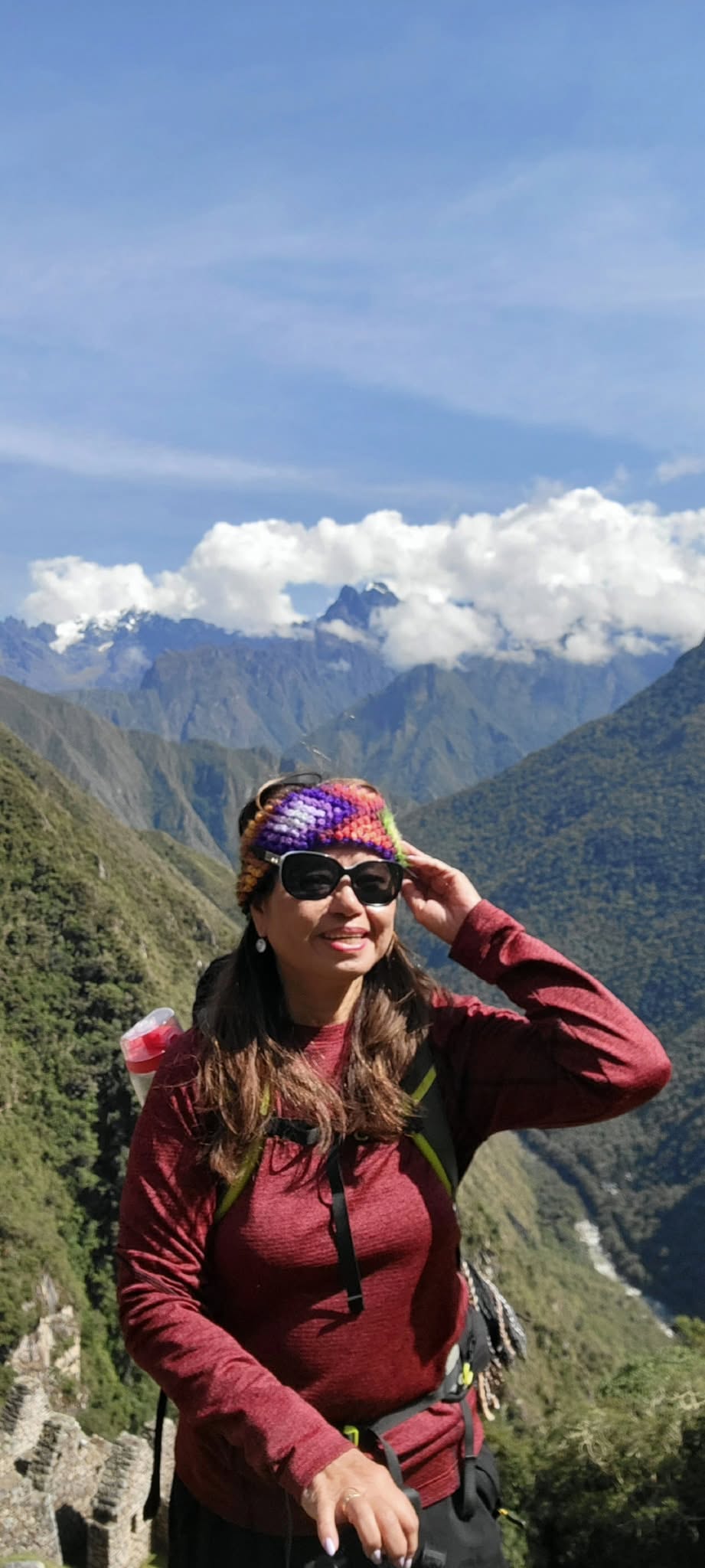 Woman in colorful knit hat and sunglasses smiling on a mountain trail with snow-capped peaks and valley in background