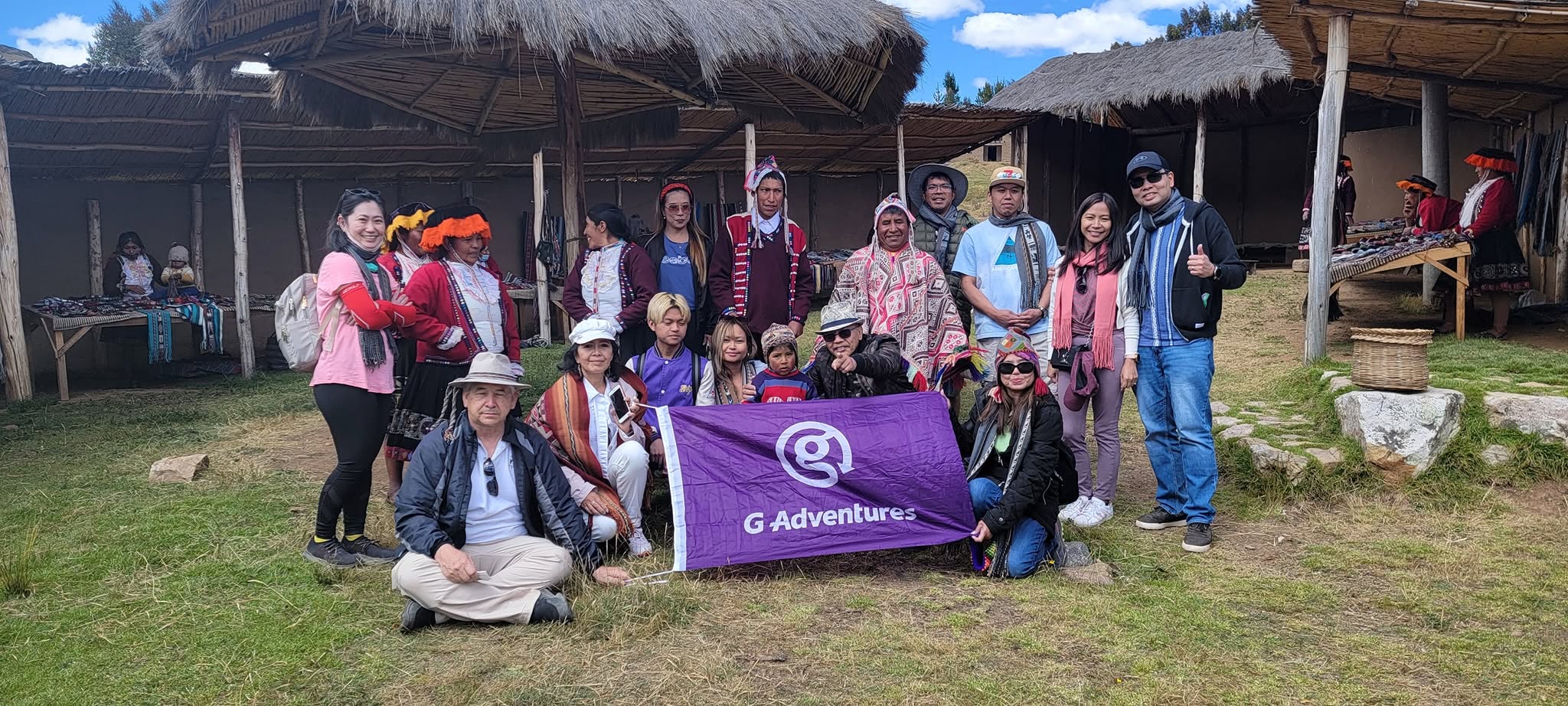 Group of tourists with G Adventures flag posing with local Peruvian guides in traditional dress at a mountain settlement