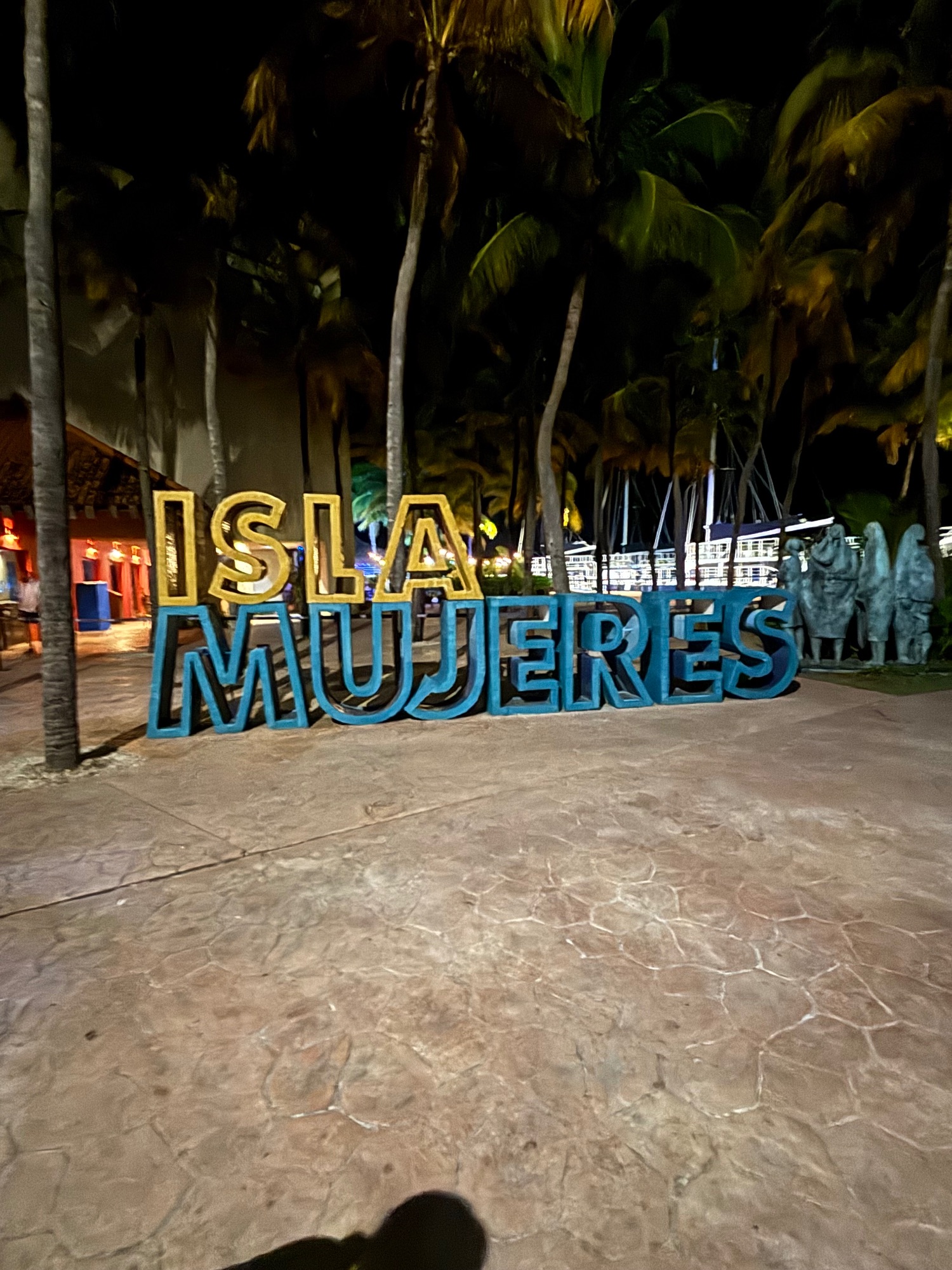 Illuminated 'Isla Mujeres' sign at night on a sandy beach with palm trees and resort buildings