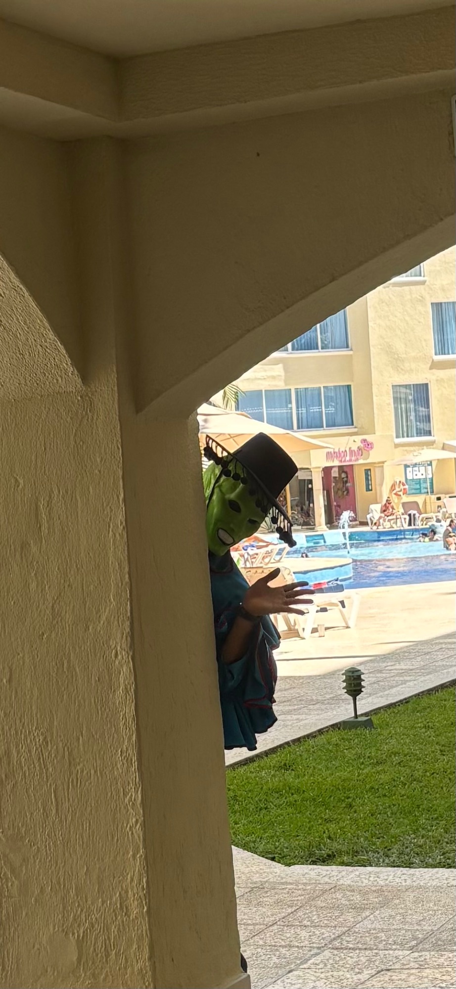 Woman in black hat peeking around a concrete pillar overlooking a resort pool and courtyard