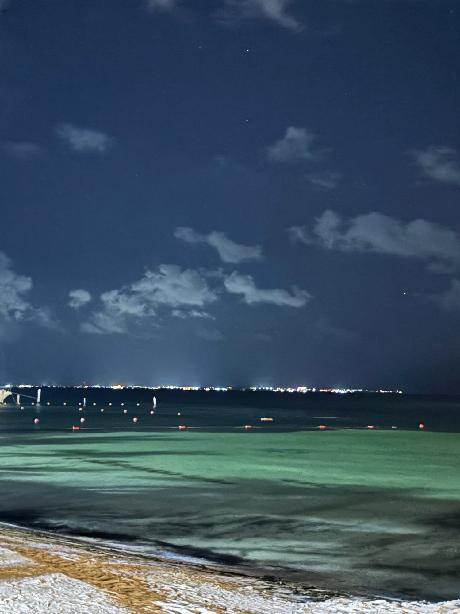 Nighttime beach view with calm turquoise water, buoys, and illuminated shoreline under dramatic sky