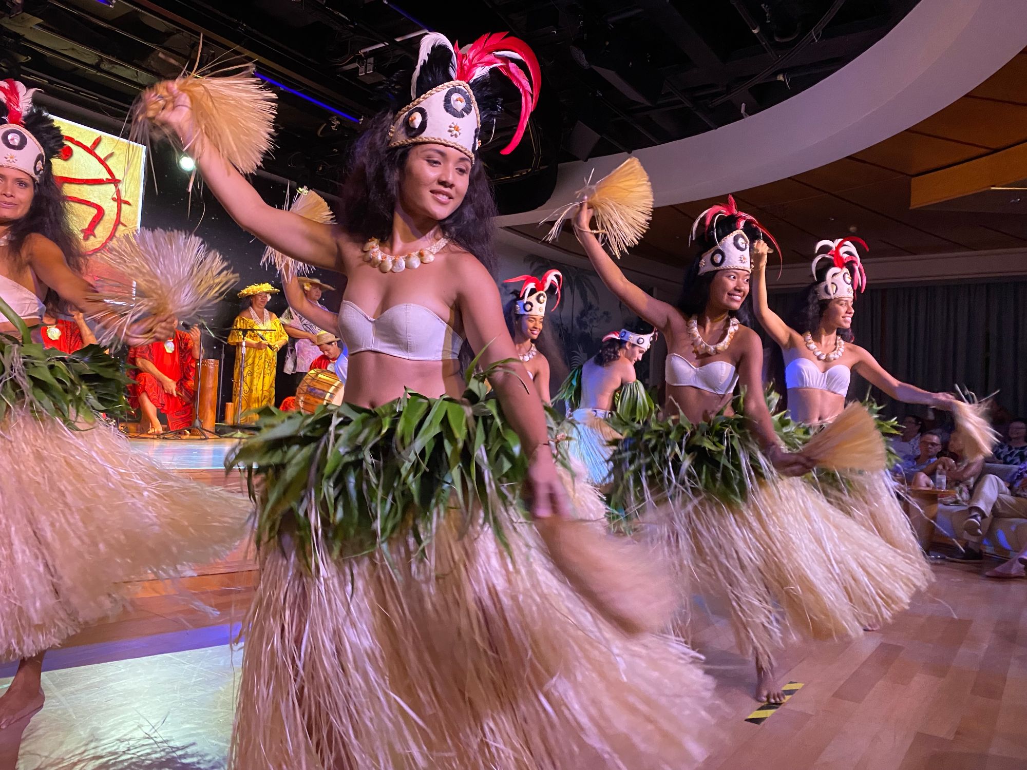 Polynesian dancers in traditional grass skirts and feathered headdresses performing on an indoor stage