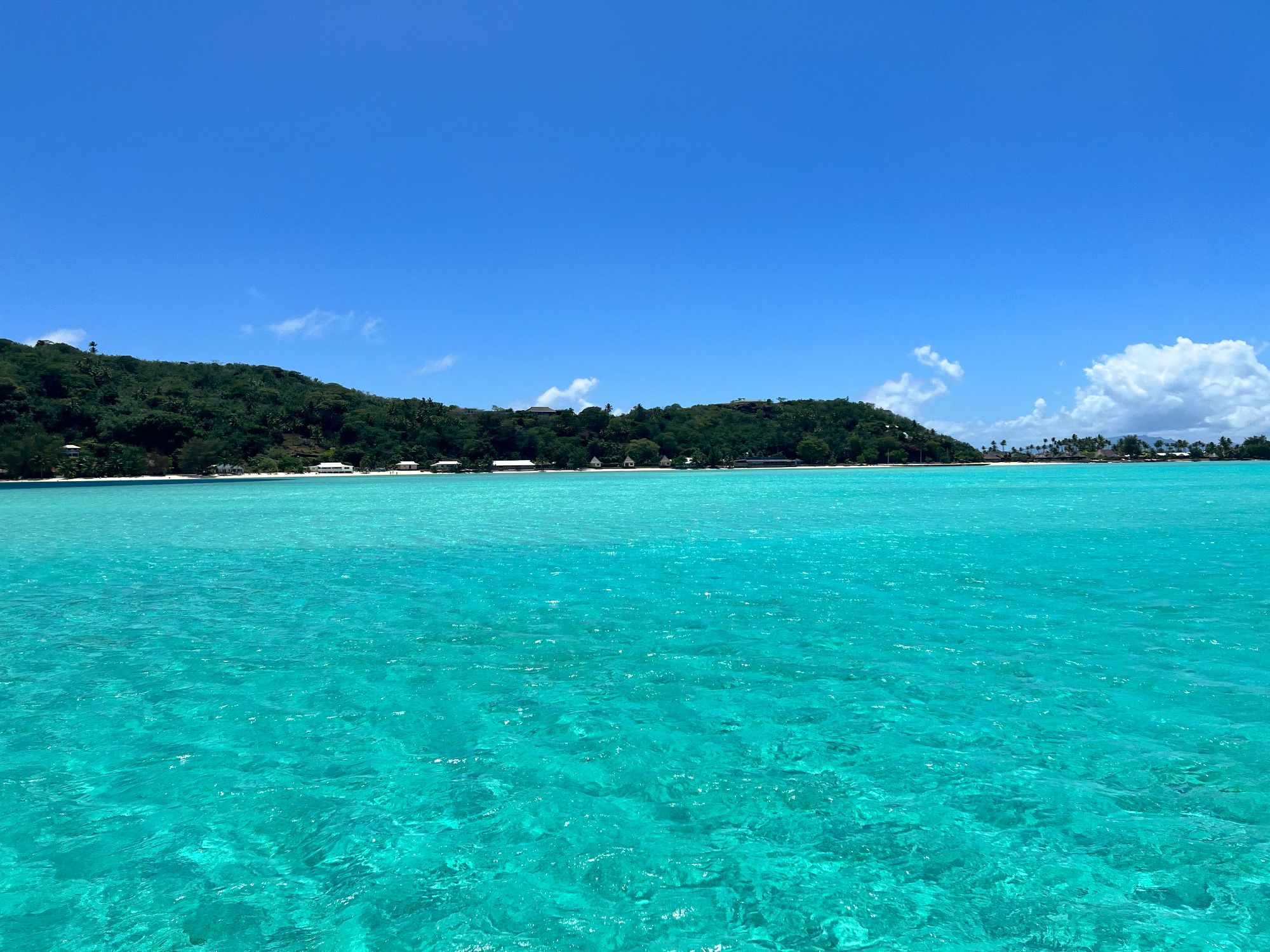 Calm turquoise lagoon with a white sand beach and lush green hillside dotted with houses along the shoreline