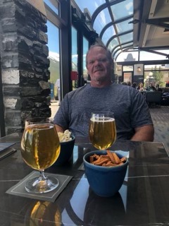 Man in a gray t-shirt smiling at an outdoor table with two glasses of beer and a bowl of appetizers