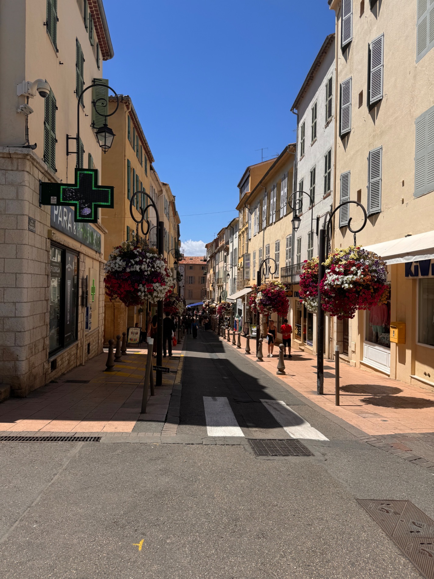 Narrow pedestrian street lined with ochre-colored buildings, hanging flower baskets, and shops in a French village