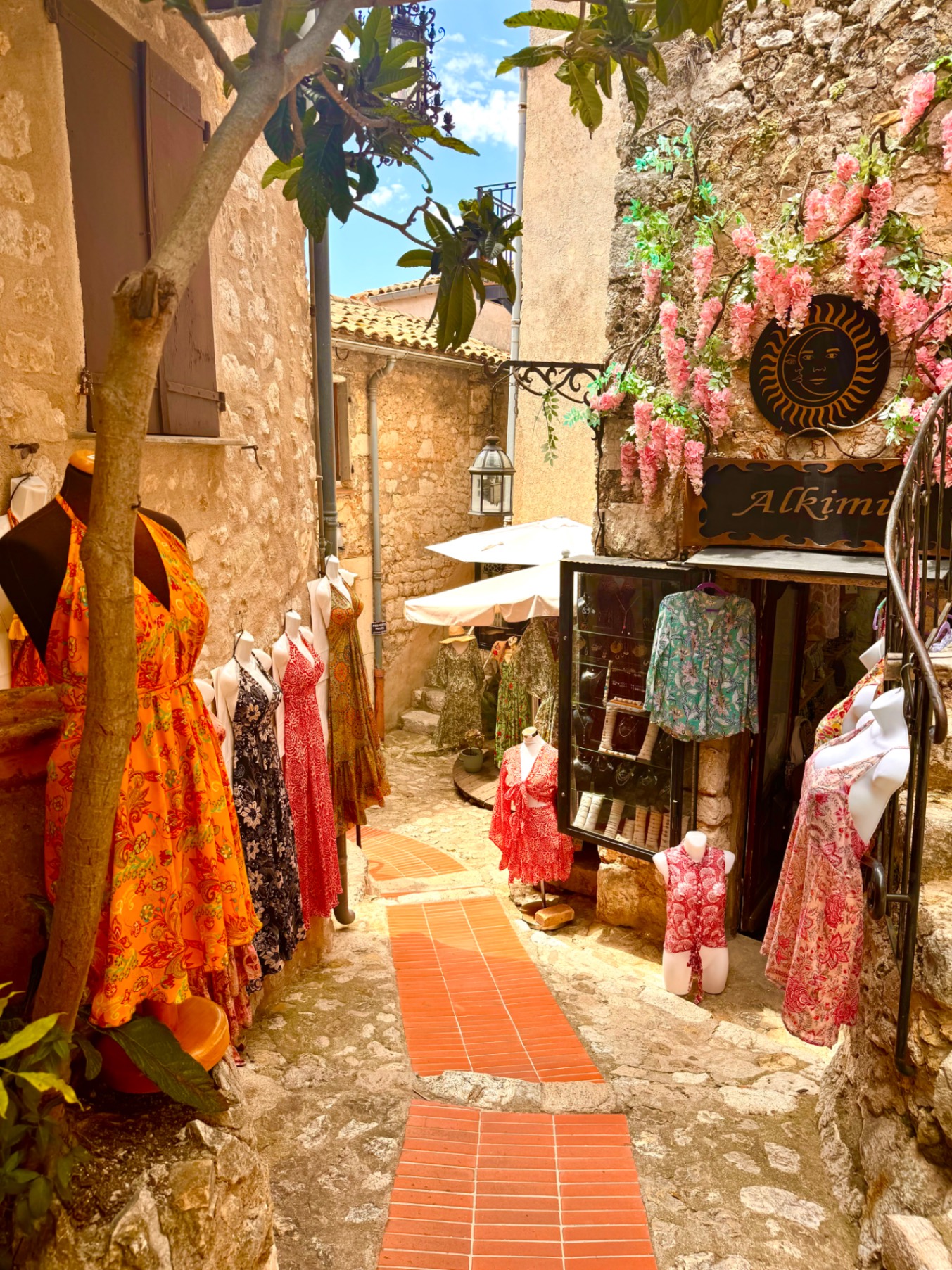 Narrow cobblestone alley with colorful dresses displayed outside boutique, pink bougainvillea, and stone buildings