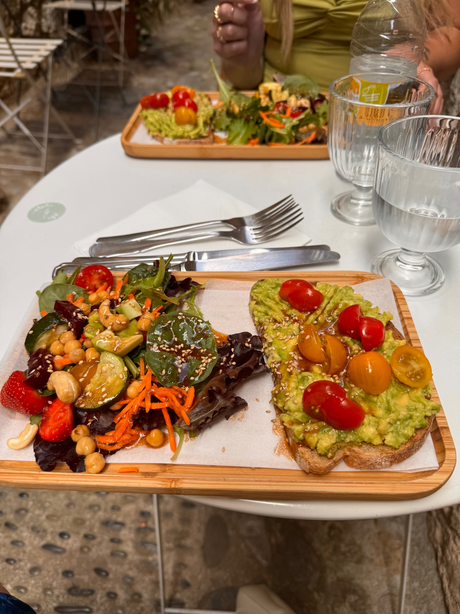 Plated avocado toast with colorful salad, cherry tomatoes, and garnishes on wooden board at outdoor café