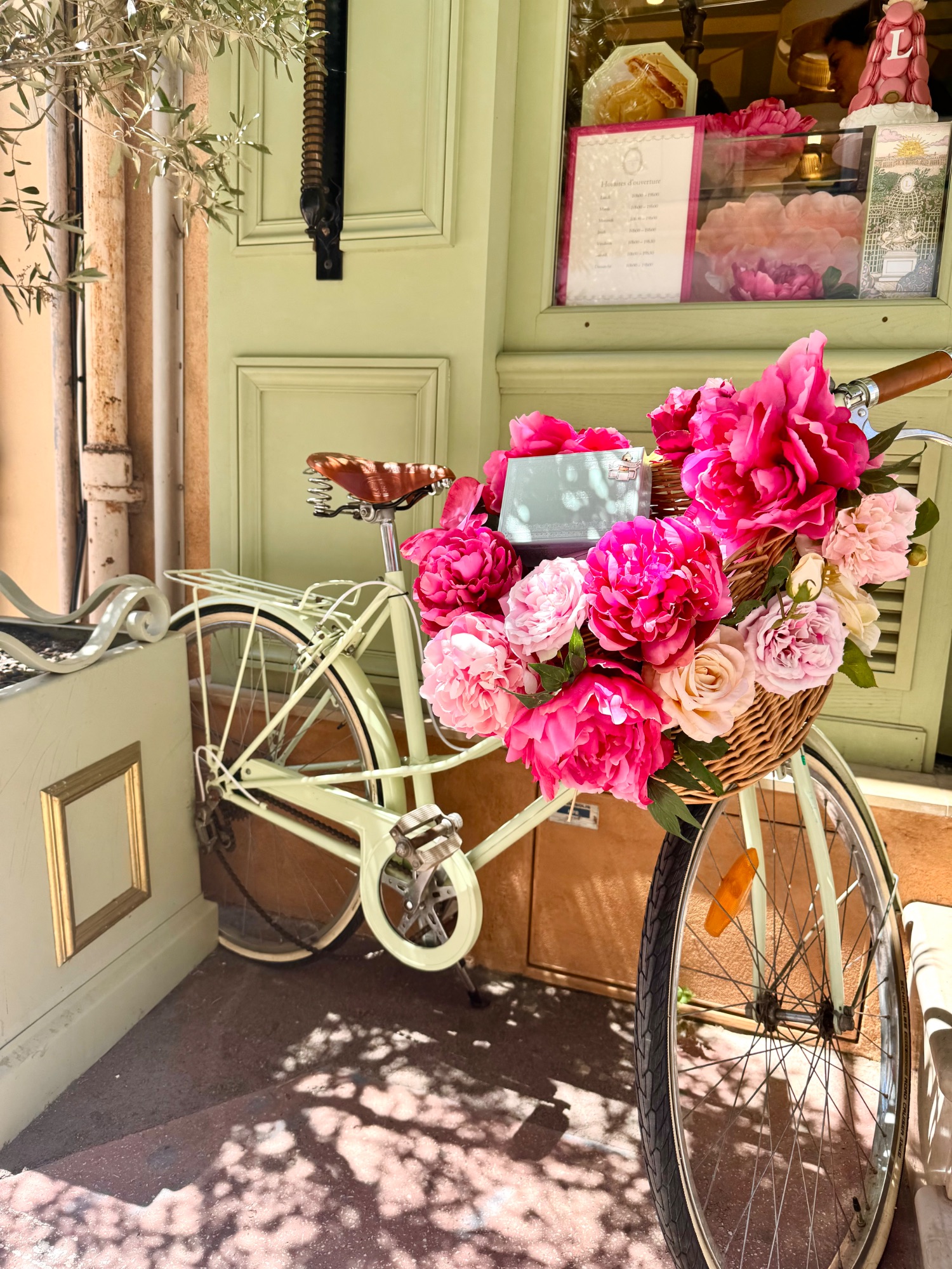 Cream-colored vintage bicycle with pink peony flowers in basket outside pale green storefront