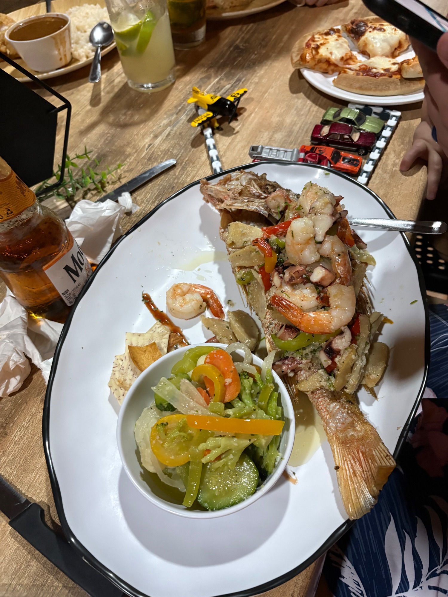 Grilled whole fish with shrimp and sautéed vegetables served on white plate with drinks