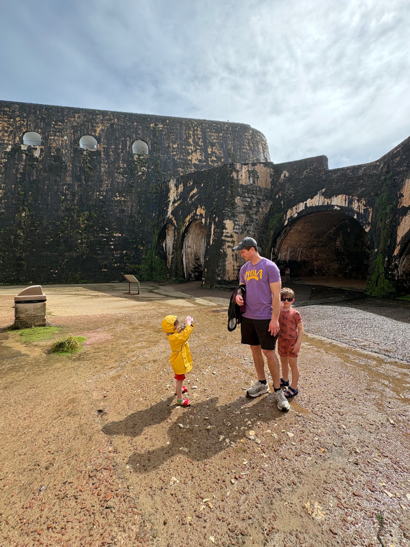 Historic stone fortress with arched openings, visitors walking in courtyard, yellow building visible
