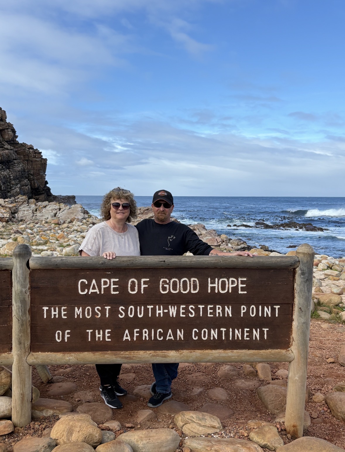 Couple posing at Cape of Good Hope sign marking the southwestern point of African continent with rocky coastline