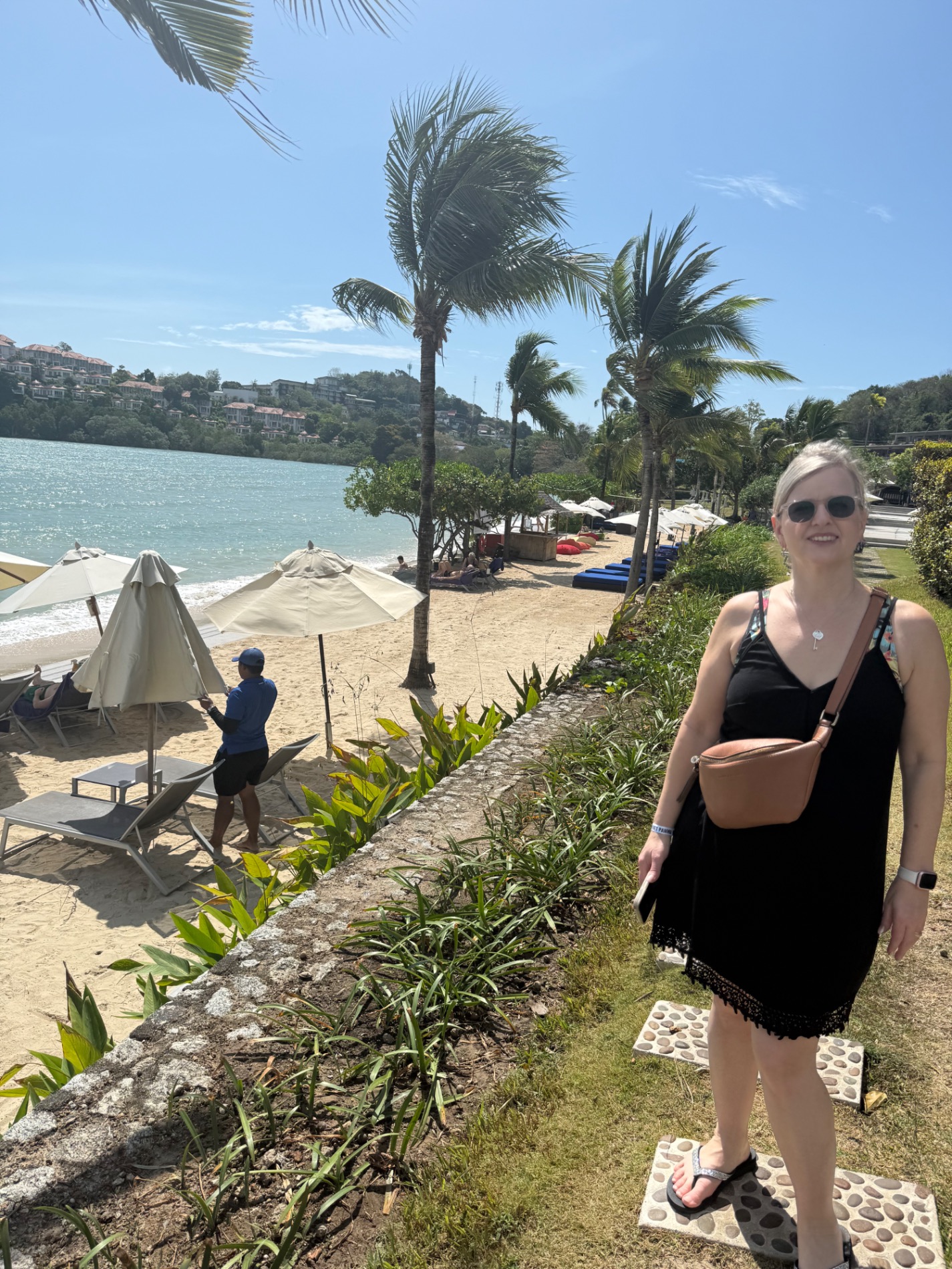 Woman in black dress standing on beach path with palm trees, umbrellas, and turquoise water visible in background